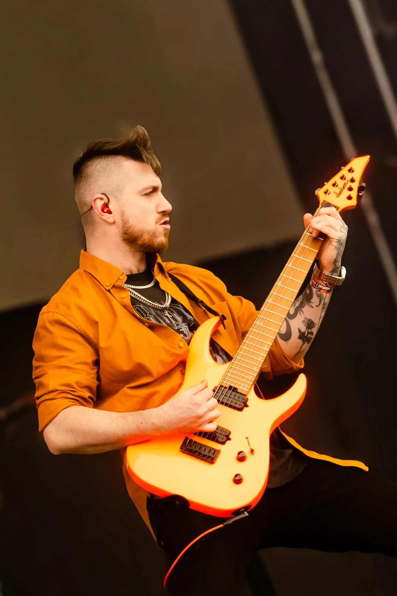 A man with tattoos, short styled hair, and a beard playing an orange electric guitar on stage, wearing an orange shirt and a black t-shirt underneath, with a focused expression and an in-ear monitor.