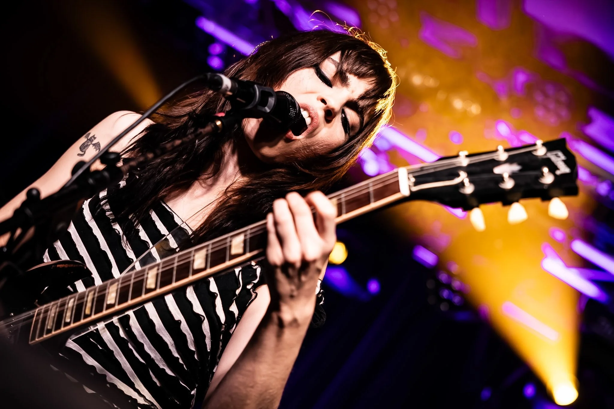 Female musician with dark hair singing into a microphone and playing an electric guitar, with colorful stage lighting in the background.