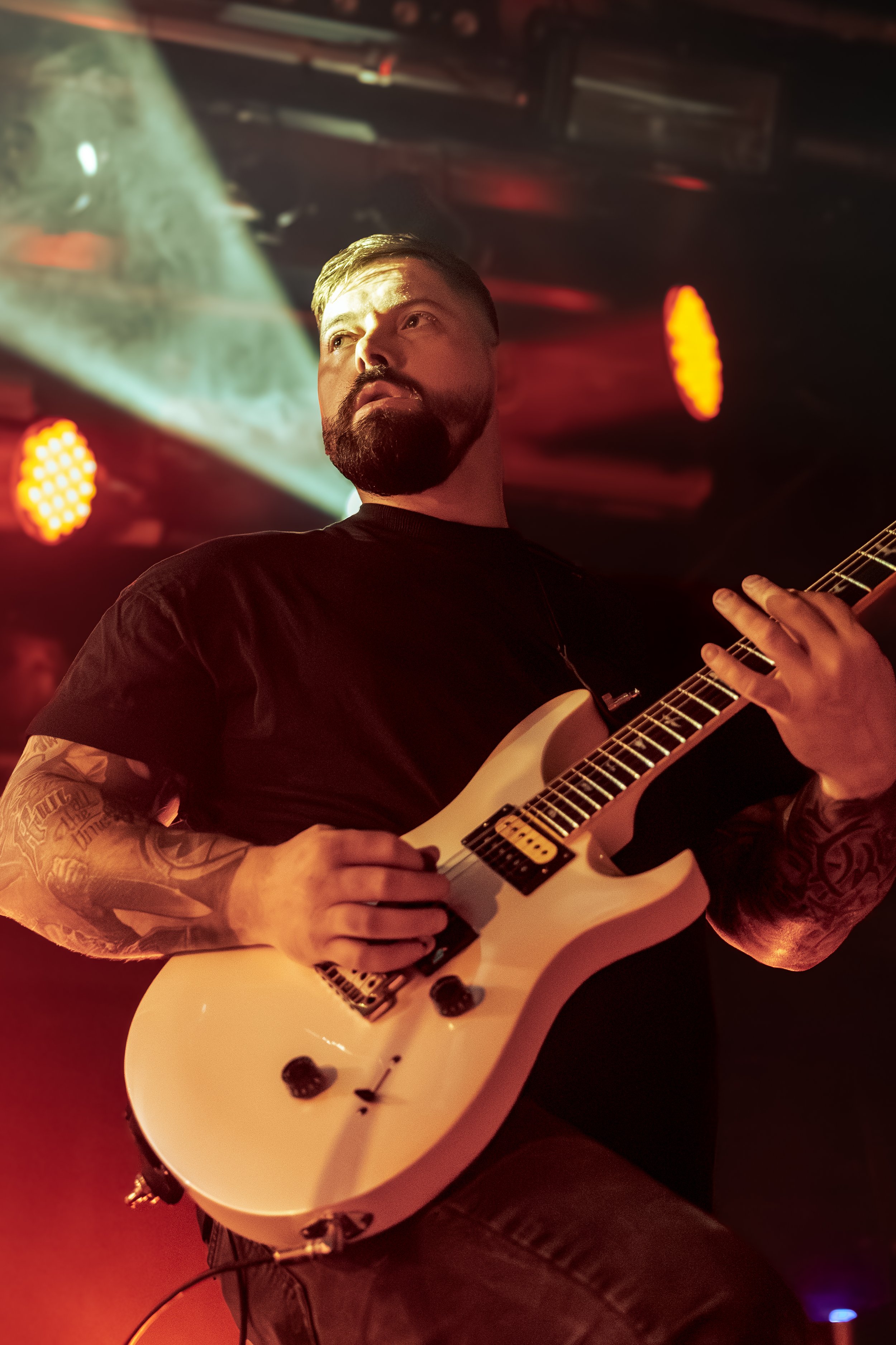 A man with a beard and tattoos playing an electric guitar on stage, with red and yellow stage lights in the background.