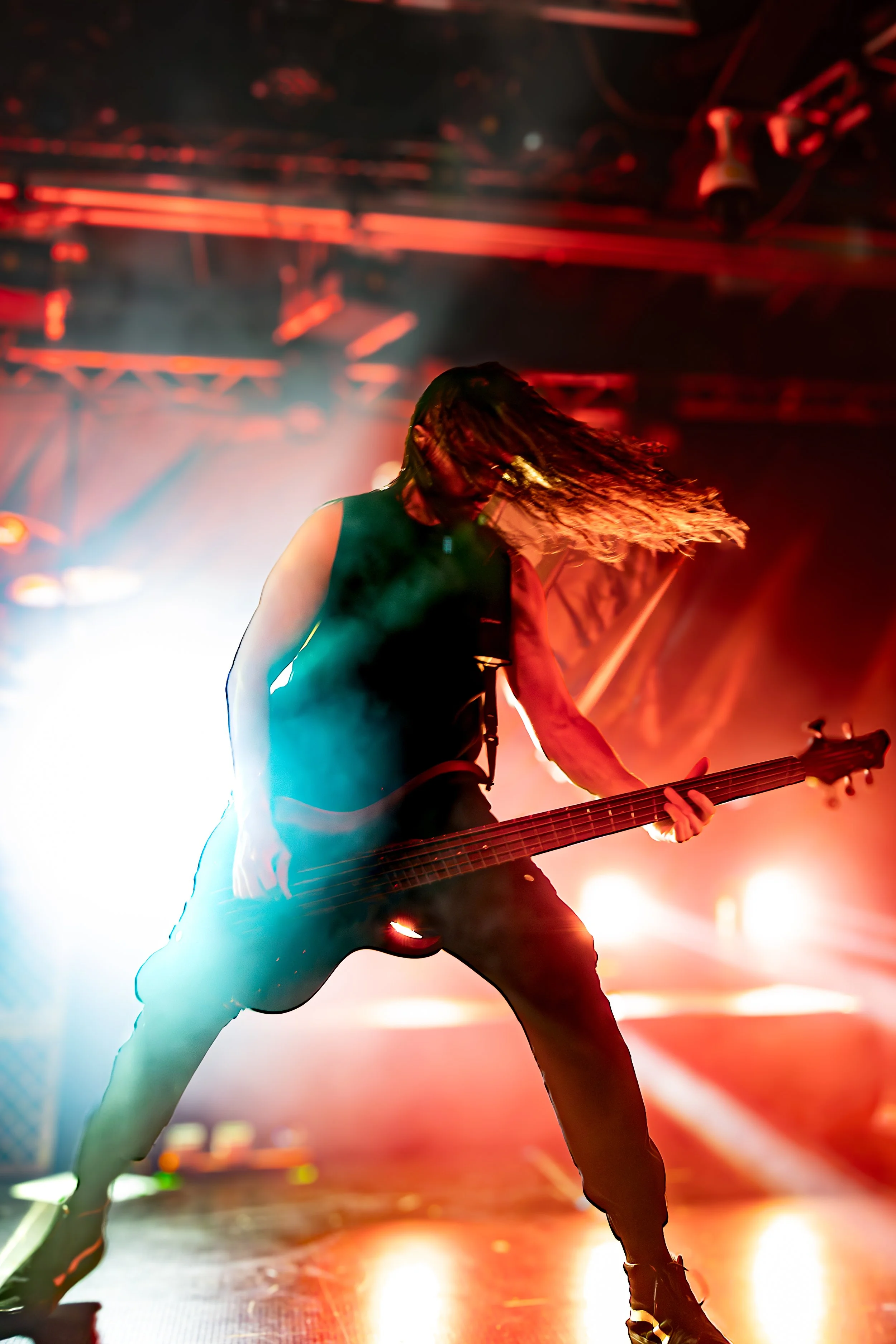 A musician with long hair playing an electric bass guitar on stage with colorful stage lights and smoke in the background.