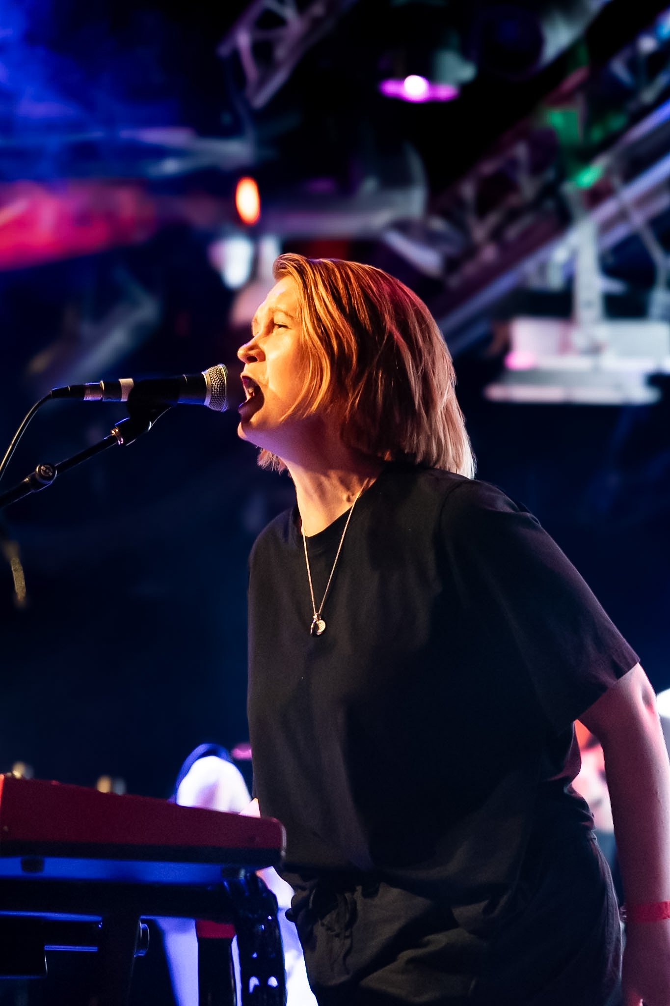 A woman with red hair singing into a microphone while playing a keyboard on stage with vibrant colorful lights in the background.