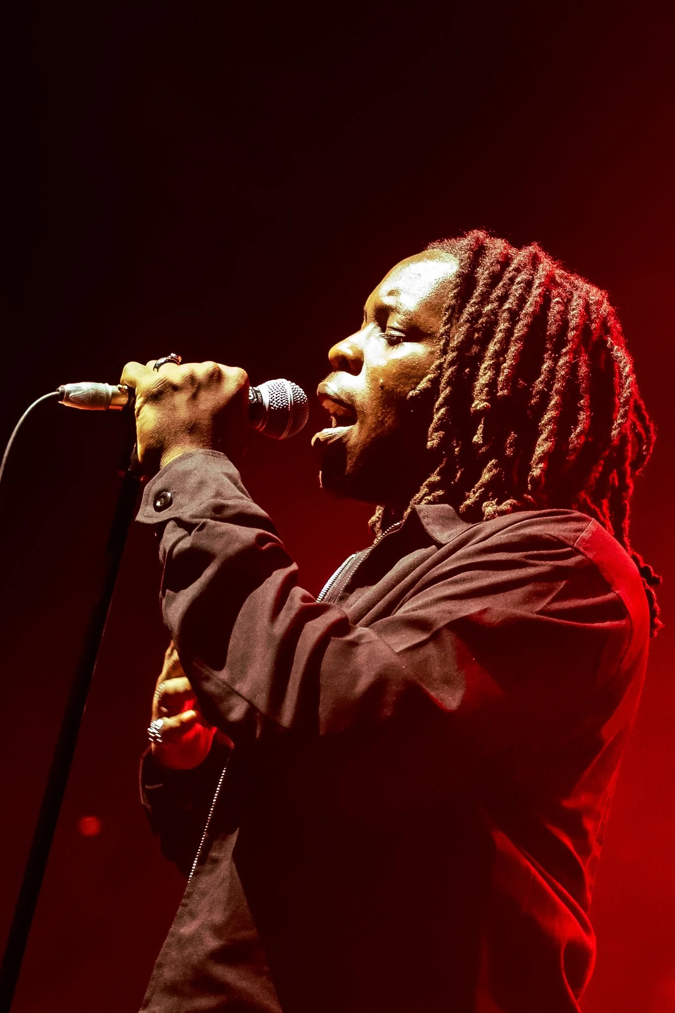 A male singer with dreadlocks holding a microphone on stage under red lighting.