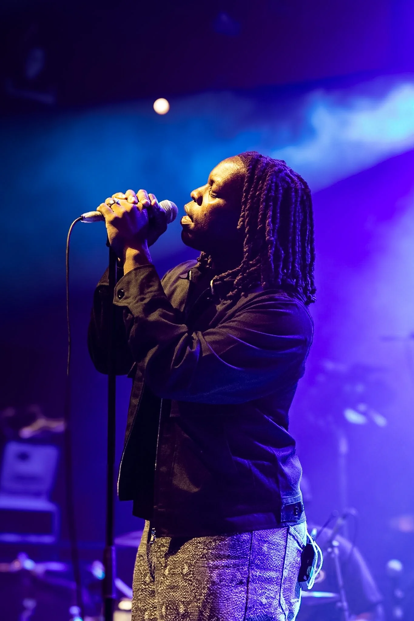A person with long braided hair singing into a microphone on stage, illuminated by purple and blue stage lighting.