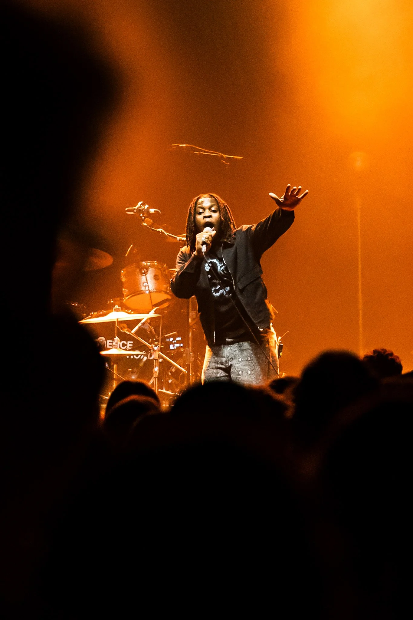 A singer with dreadlocks wearing a black jacket performing on stage, with drums and musical equipment in the background, bathed in warm orange lighting.