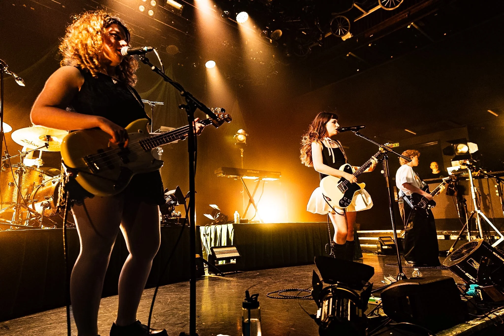 Two female musicians performing on stage under warm stage lighting. One with curly hair playing bass guitar, the other with straight hair playing an electric guitar. Both are singing into microphones, with drums and additional musical equipment in th
