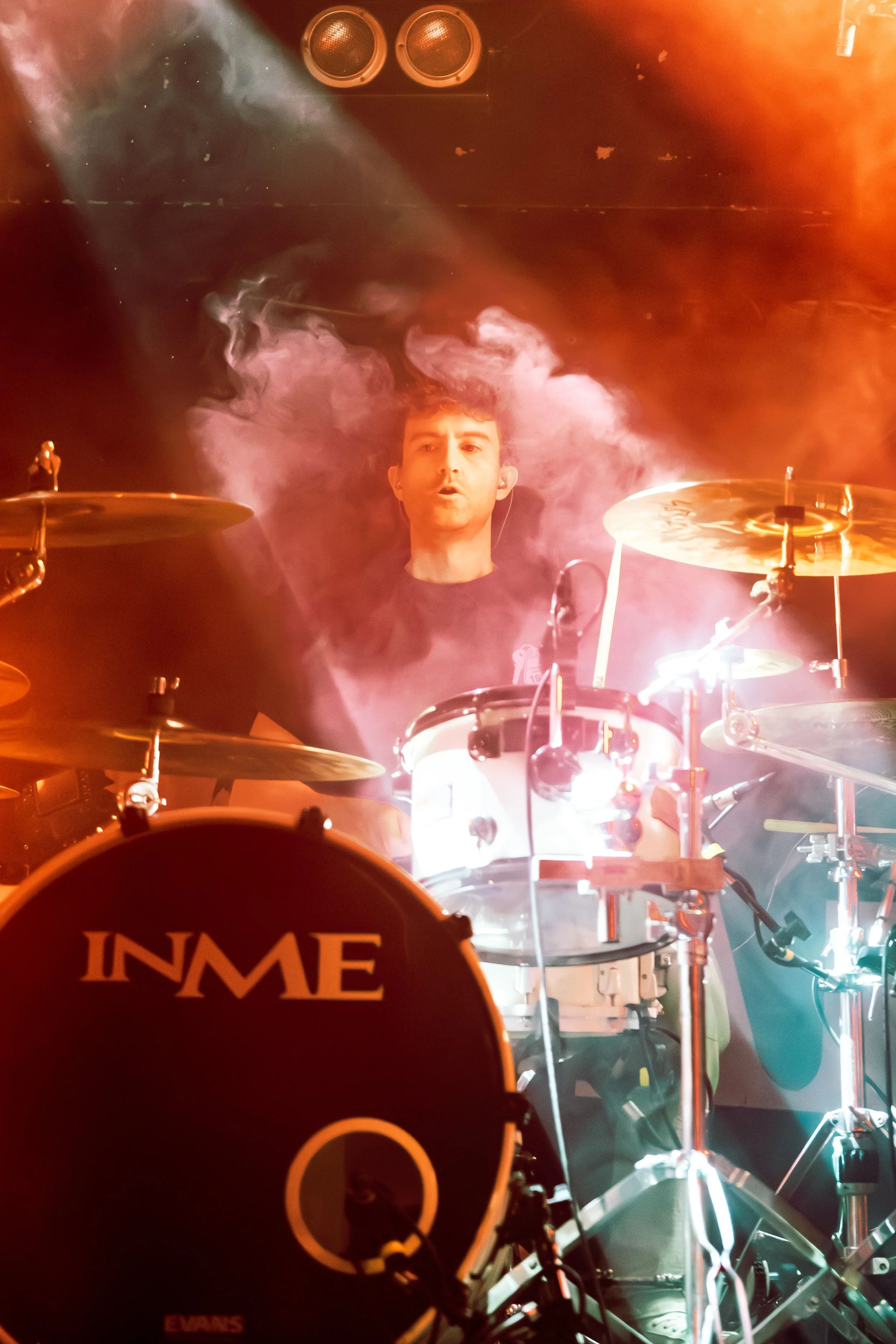 A drummer performing on stage with smoke effects and stage lighting. The focus is on the drummer's face and drum set.