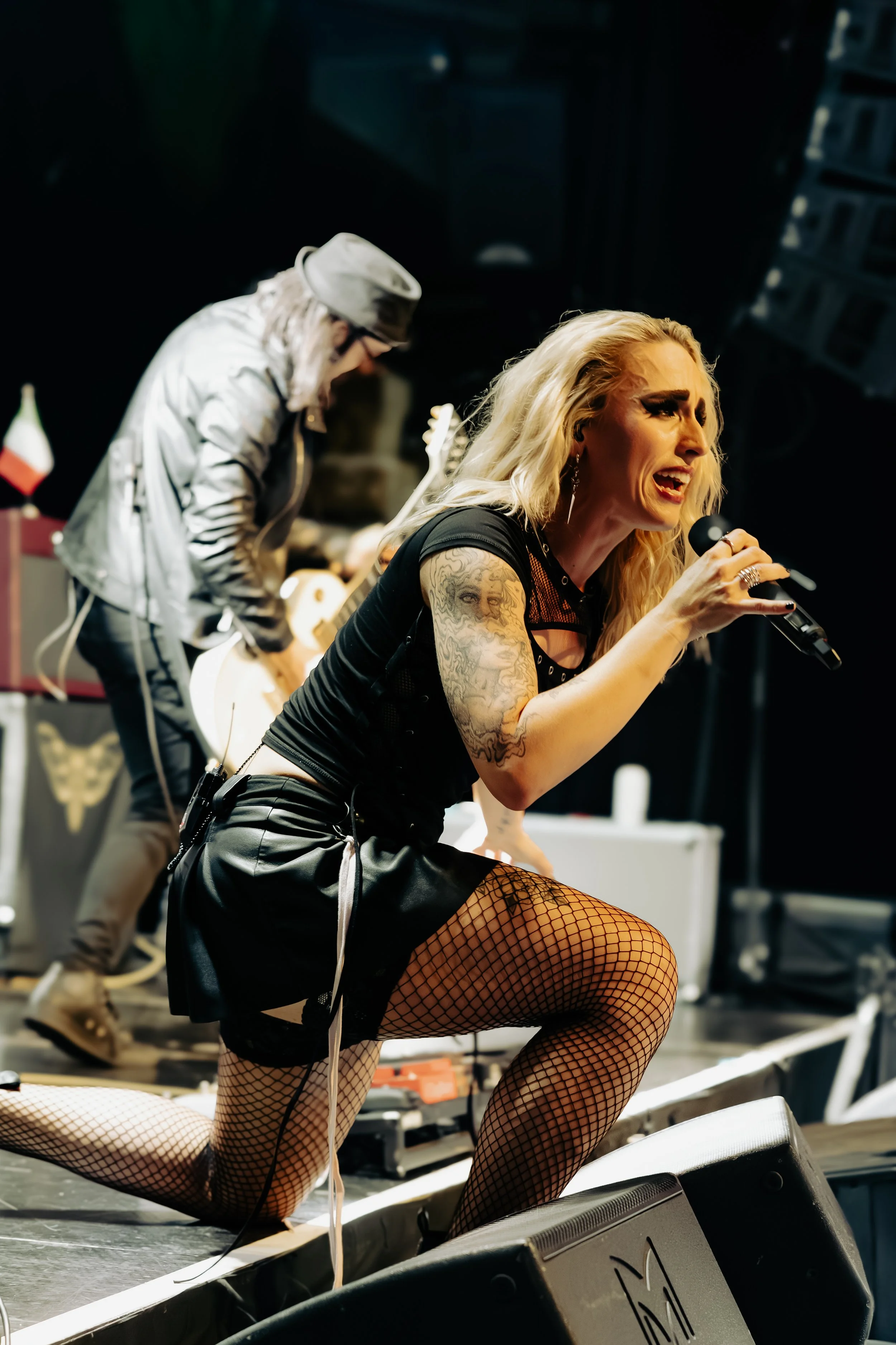 A woman with blonde hair, tattoos, fishnet stockings, and wearing black shorts, singing into a microphone on stage with a guitarist in the background.