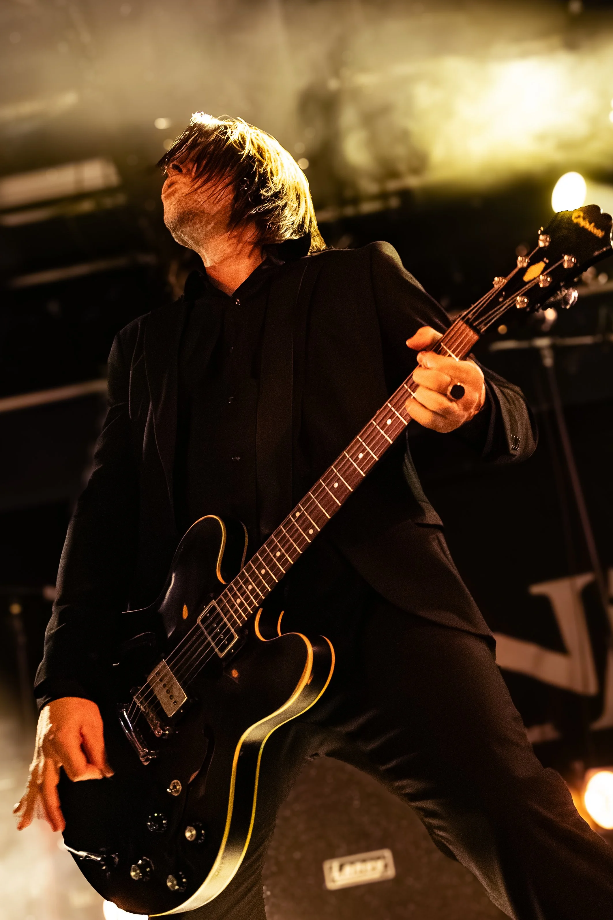 A male musician playing an electric guitar on stage, with hair covering his face, dressed in black, under warm stage lighting.