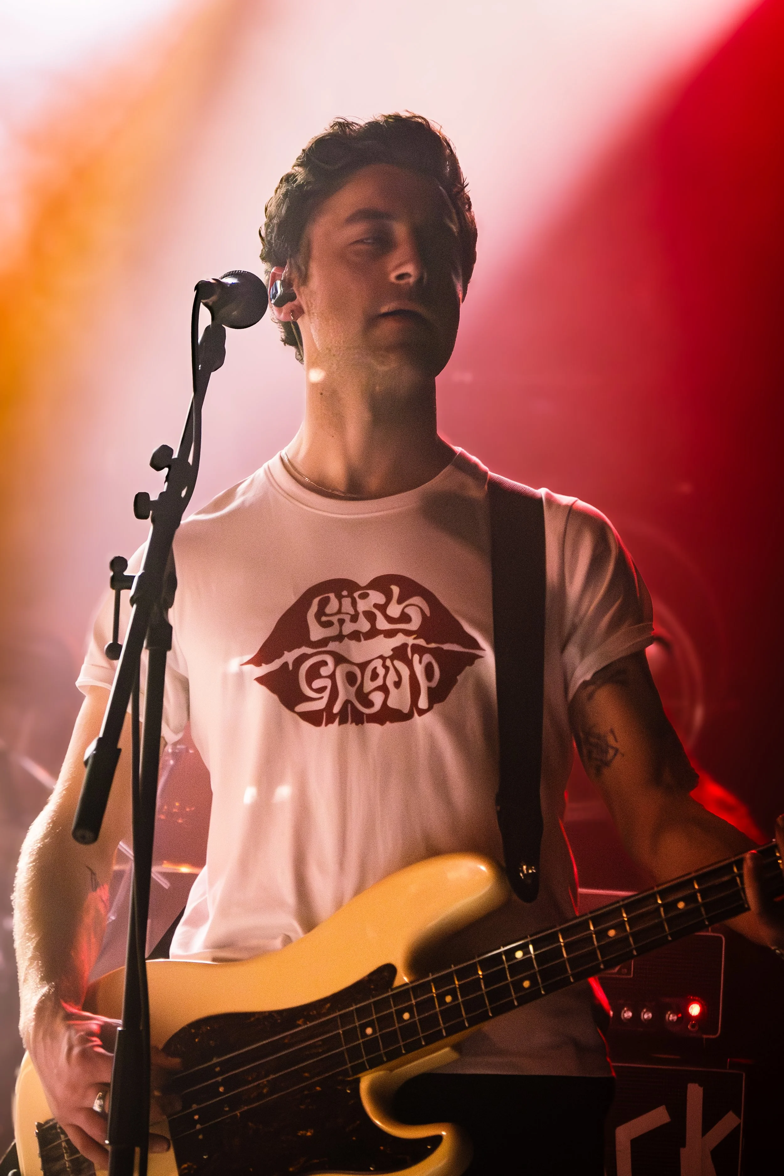 A person with dark hair playing an electric guitar on stage, wearing a white T-shirt with a graphic design, in front of colorful stage lighting.