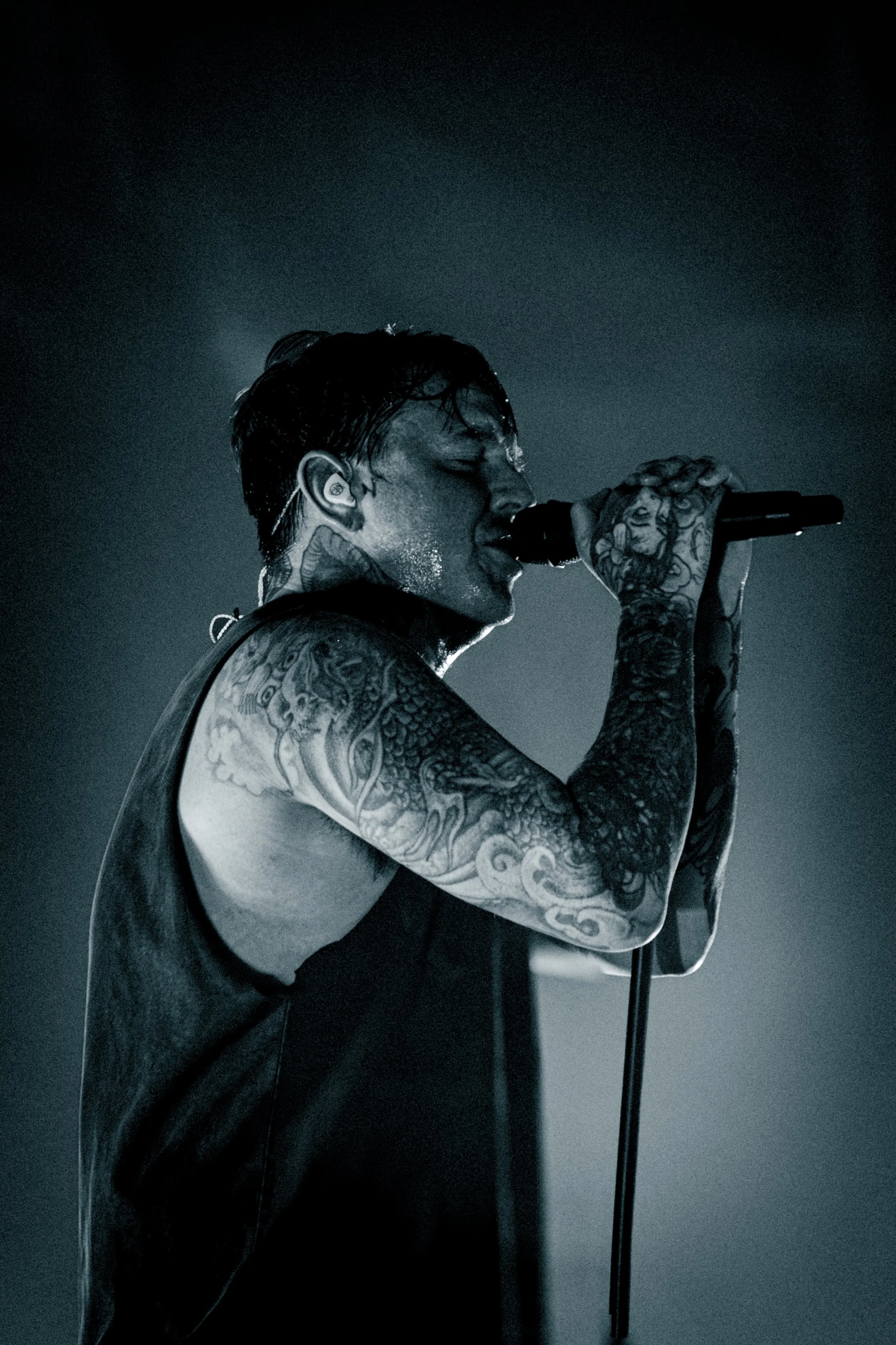 A male singer with tattoos on his arms and neck, holding a microphone close to his mouth, performing in a dark setting with a faint light illuminating his face and body.