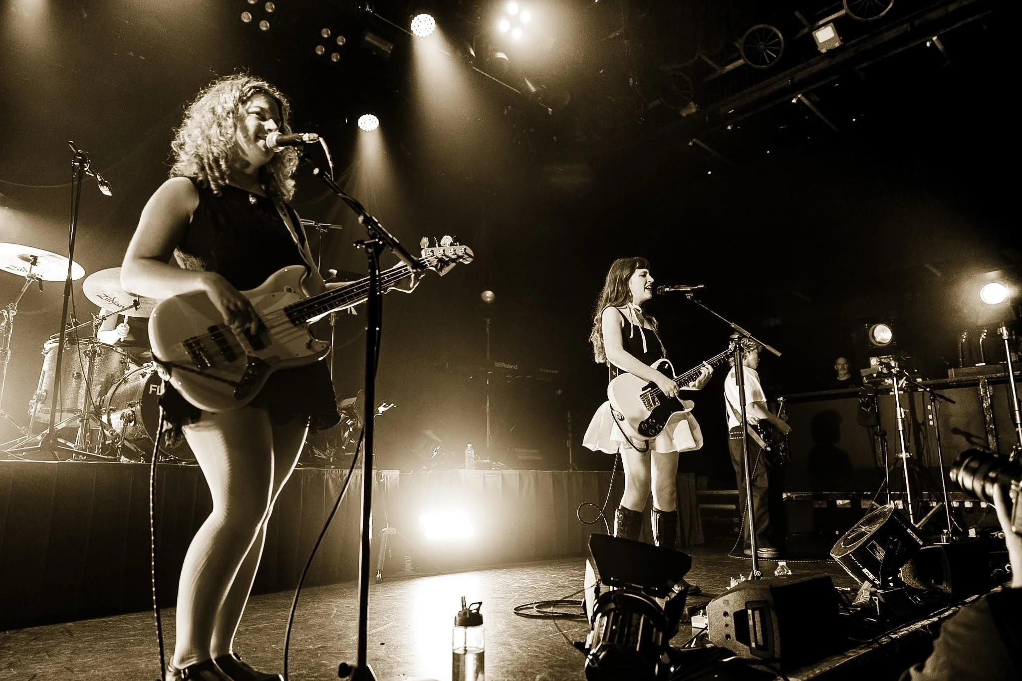 Female musicians performing on stage with guitars and microphone, bright stage lights, and musical equipment visible in the background.