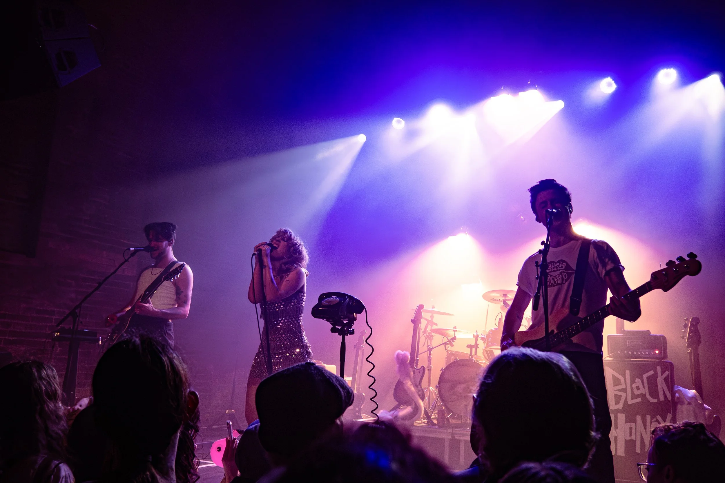 A band performs on stage under purple and blue stage lighting with female and male singers and musicians, audience members visible in the foreground.