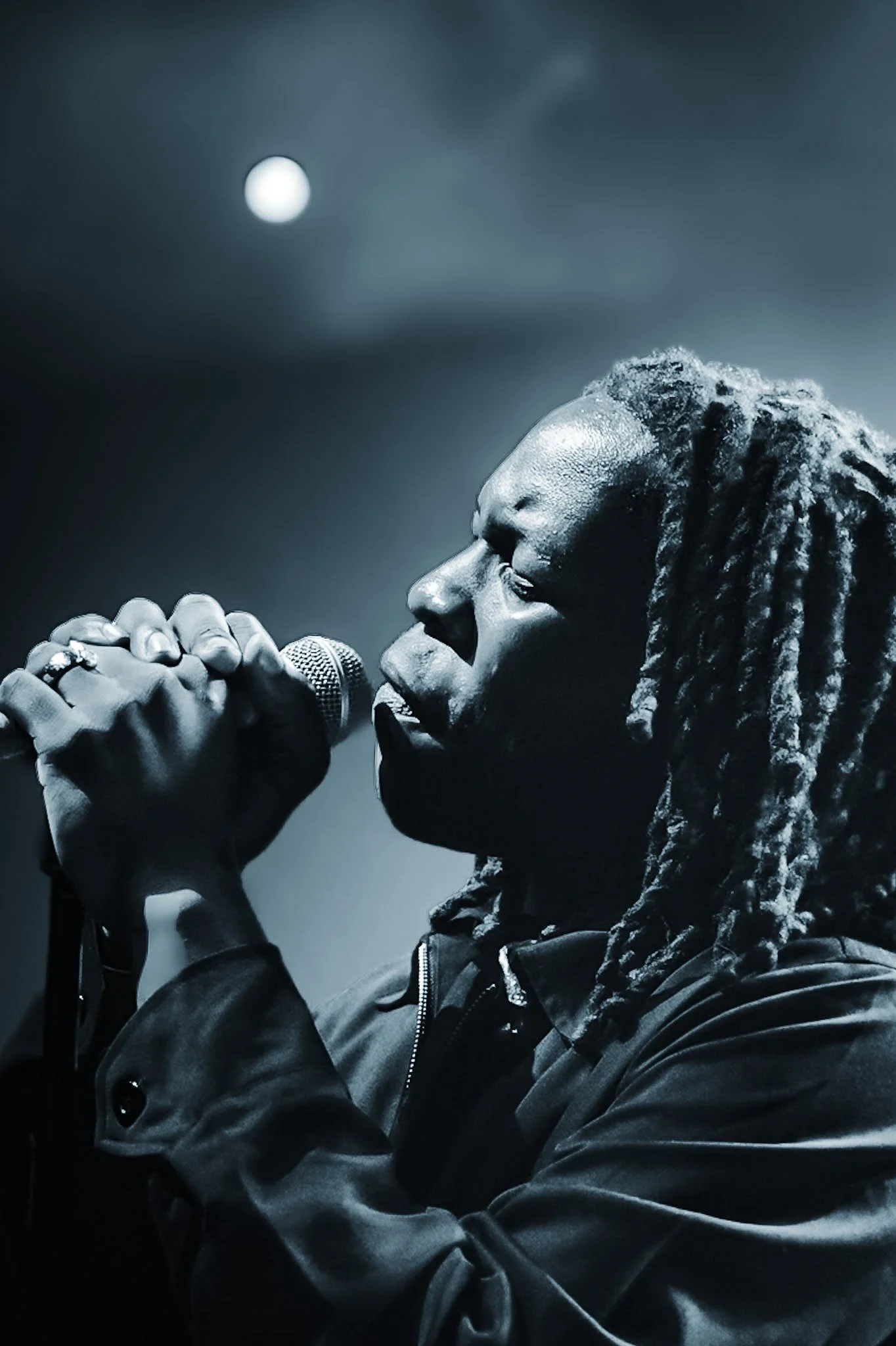 A person with long dreadlocks singing into a microphone, wearing a dark hoodie, with moonlight and cloudy night sky in the background.