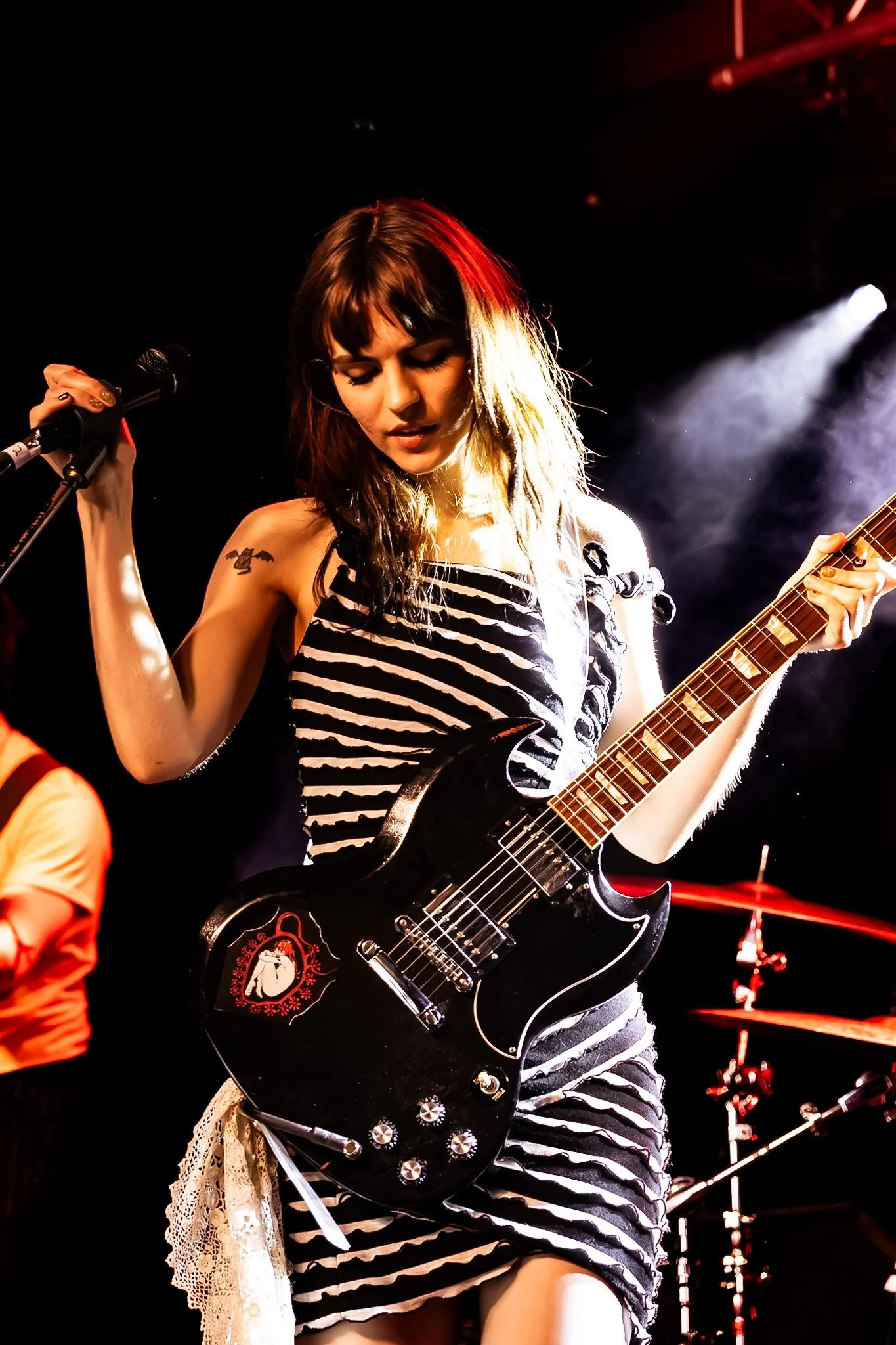 A female musician with brown hair and bangs playing an electric guitar on stage. She is wearing a black and white striped dress and has a tattoo on her right shoulder. The background is dark with stage lights.