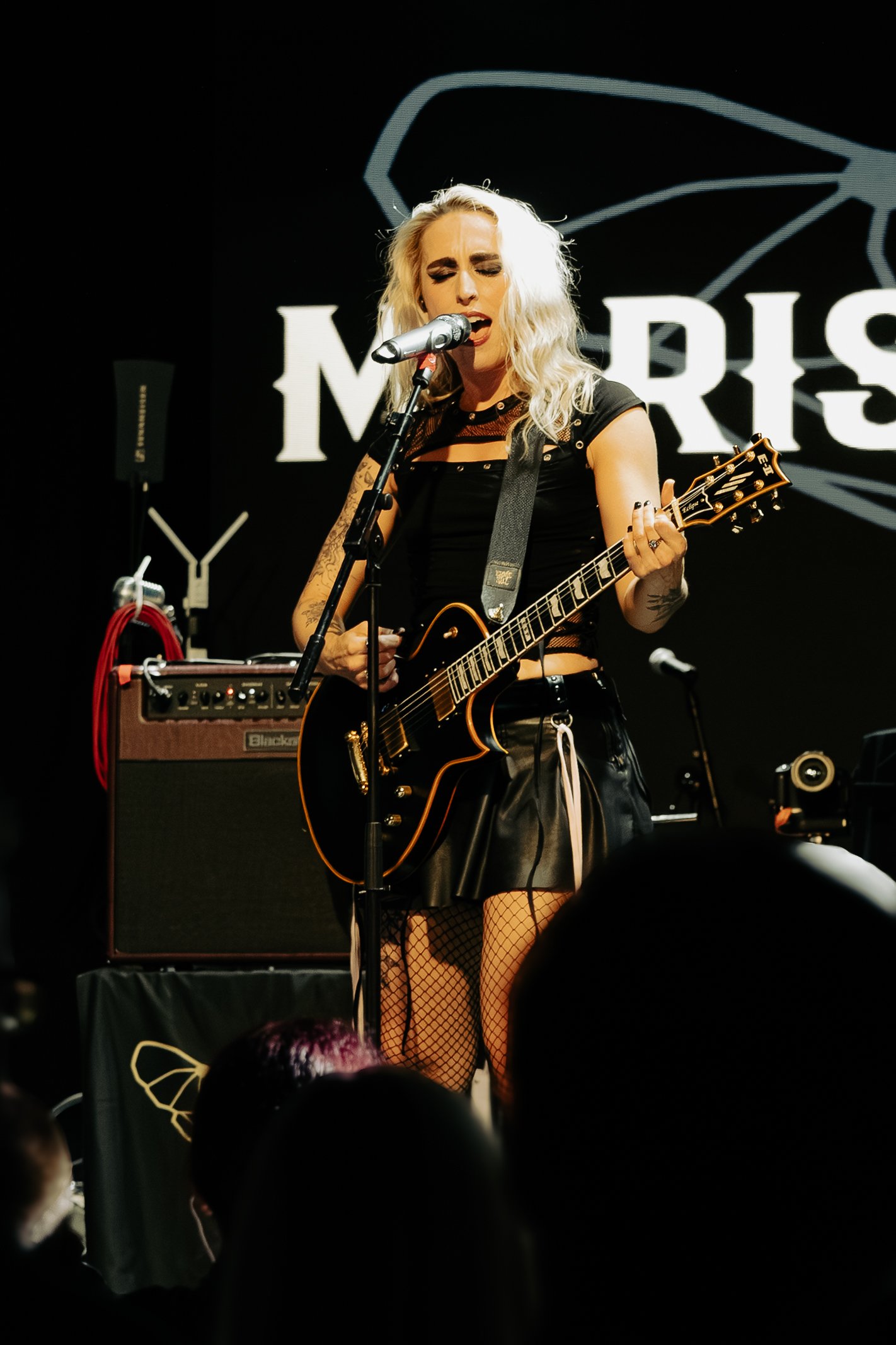A woman with blonde hair singing into a microphone while playing guitar on stage. She is wearing a black outfit with fishnet stockings and appears to be performing passionately. Behind her, part of a logo or sign is visible on a dark background.