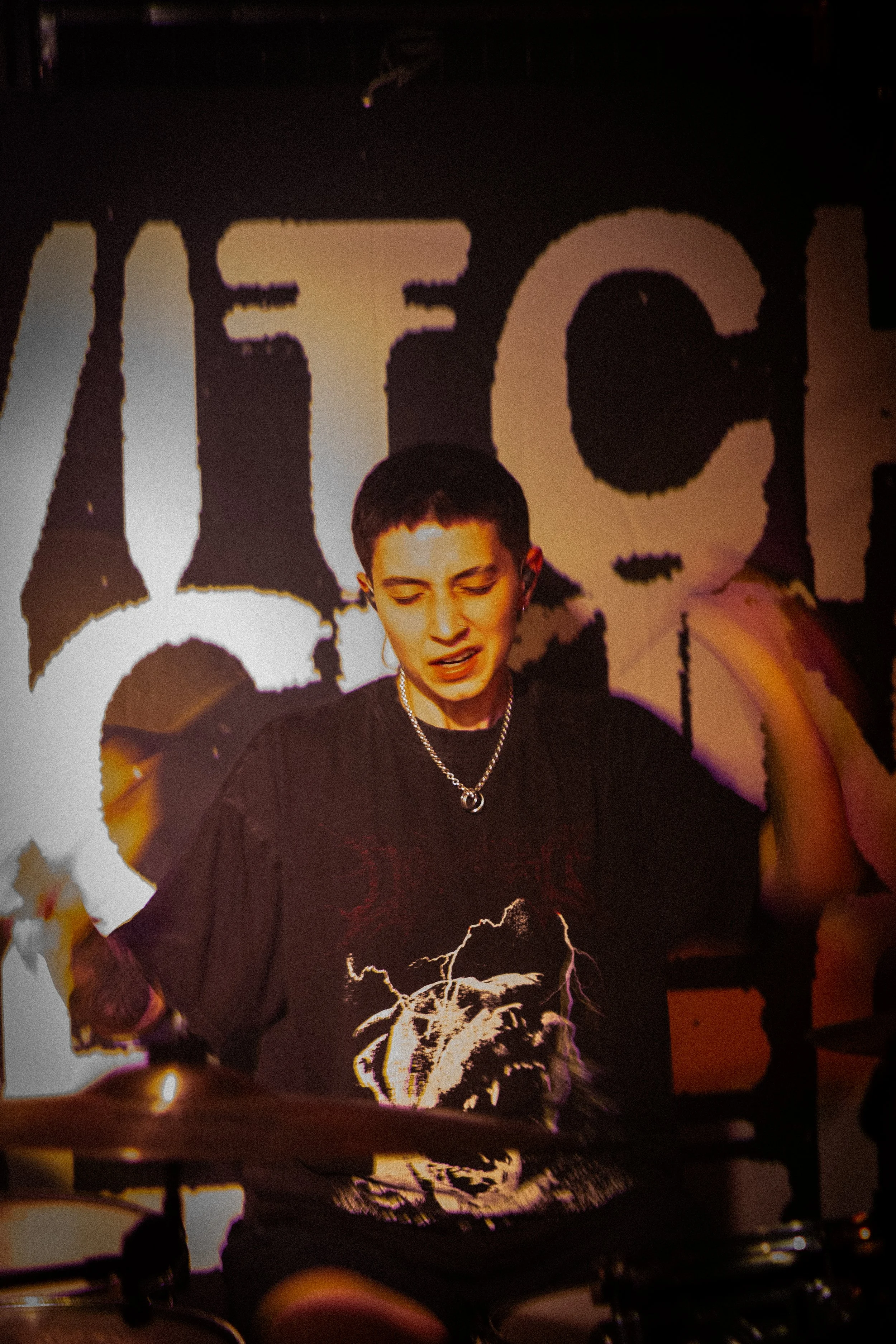 A person with short dark hair and light skin, wearing a black shirt with a white graphic design of a roaring lion, is playing drums in front of a background with the word 'MET' in large letters.