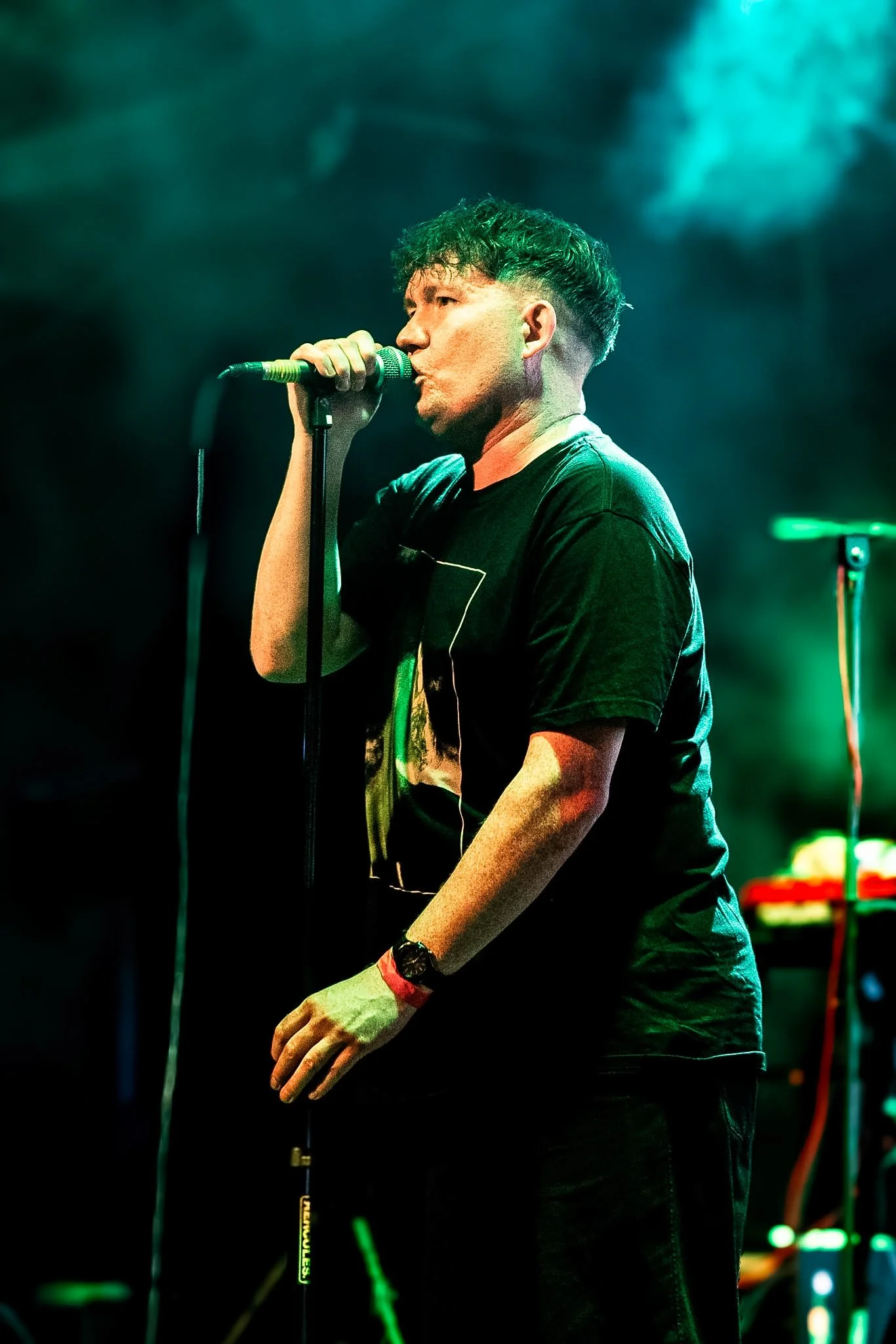 A young man with short, curly hair singing into a microphone on stage, wearing a black T-shirt and a wristwatch, with colorful stage lighting.