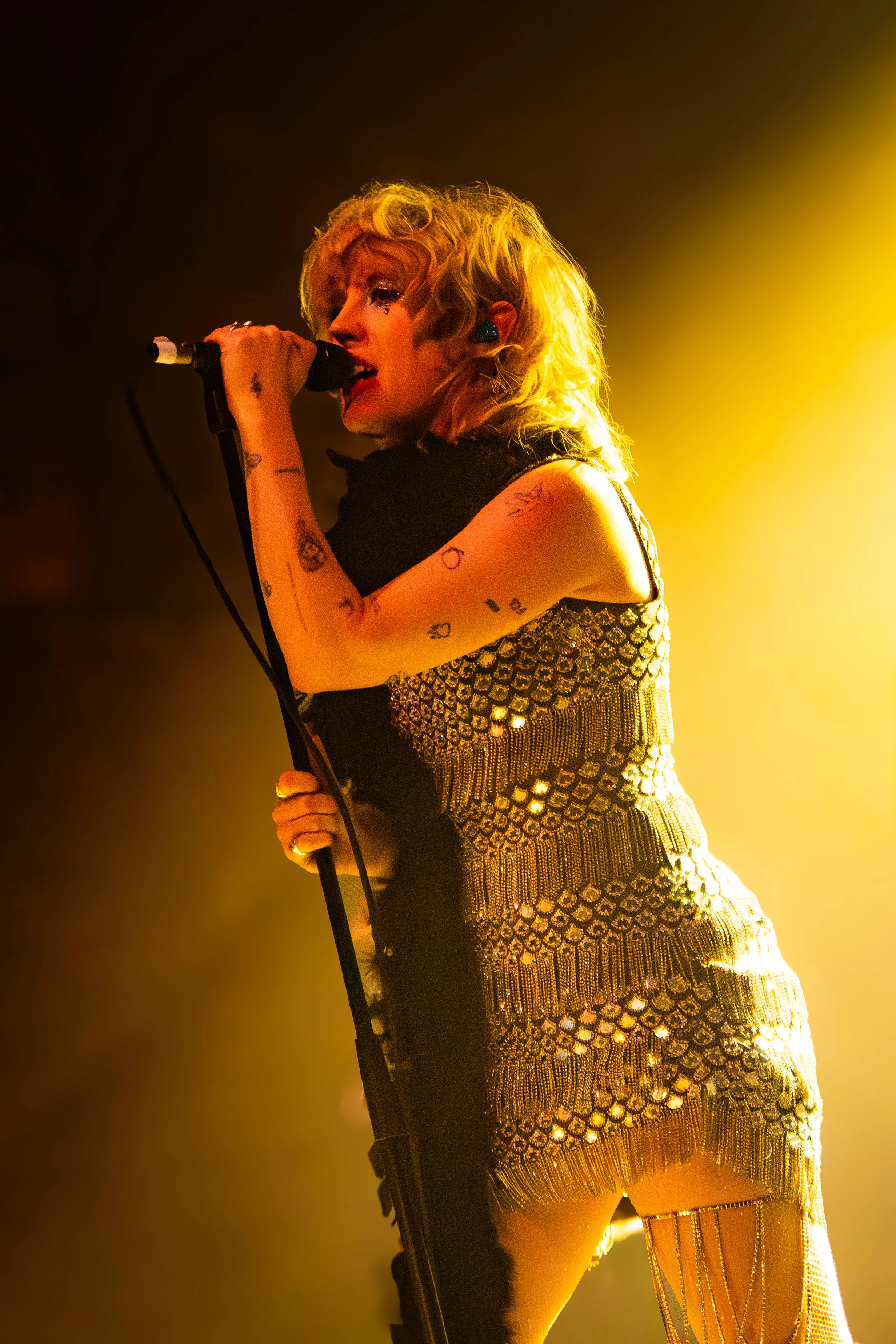 A female singer performing on stage, holding a microphone, dressed in a glittering, gold and black outfit, with blonde curly hair, illuminated by yellow stage lights. Black Honey in concert at Village Underground, London