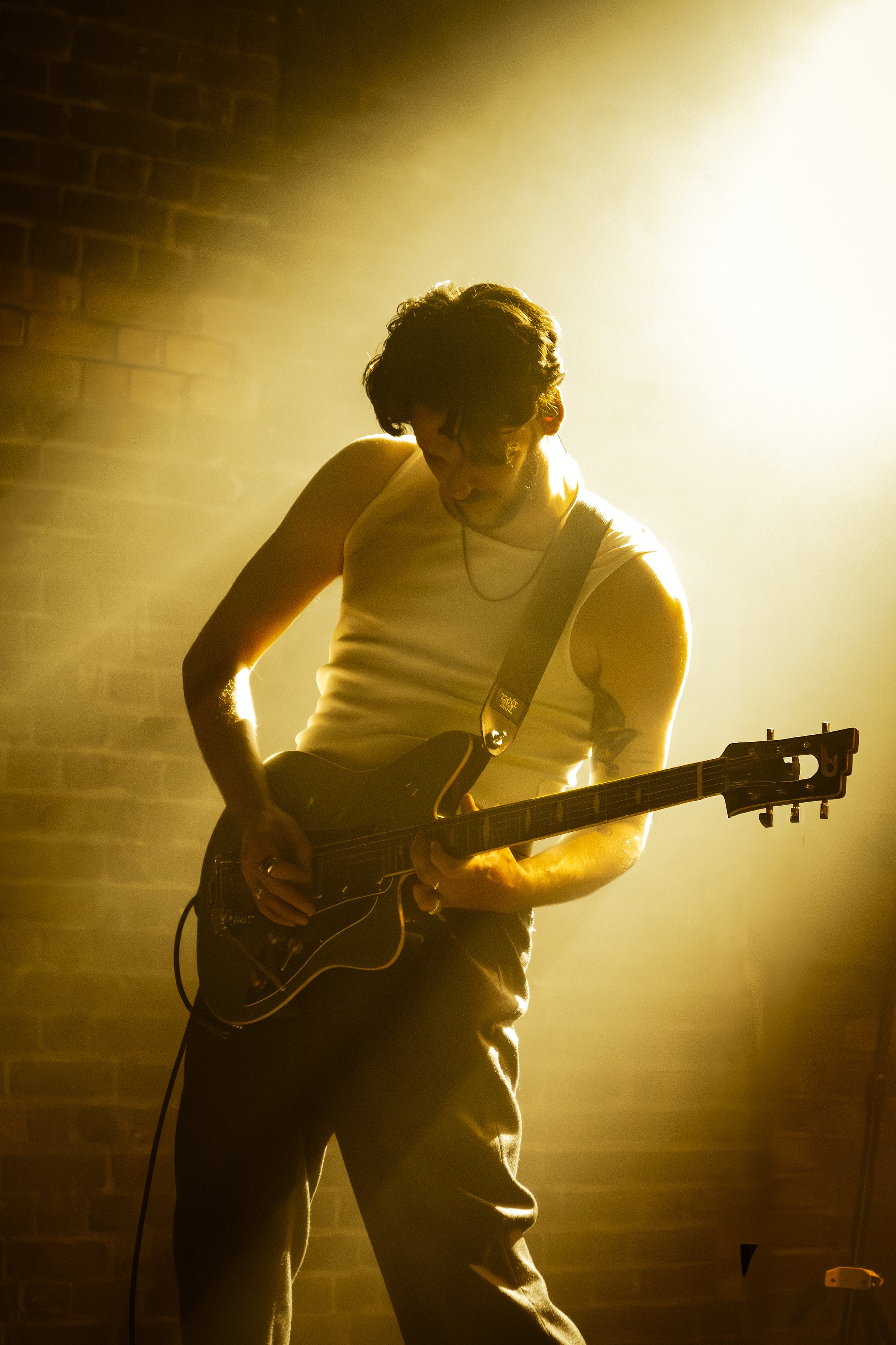 A person playing an electric guitar on stage, backlit by bright yellow light, with a brick wall in the background.