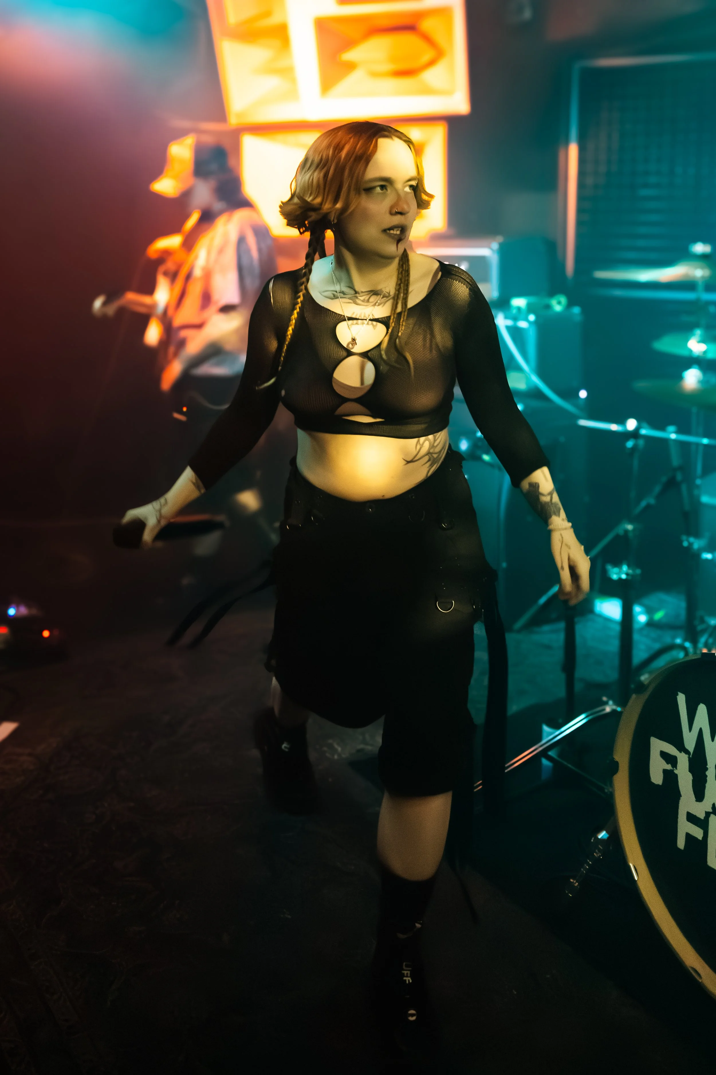 A young woman with red hair, wearing a sheer black long-sleeve top with cutouts, black shorts, and thigh-high stockings, standing under colorful neon lights in a bar or club setting. She has tattoos and wears necklaces. In the background, there is a 