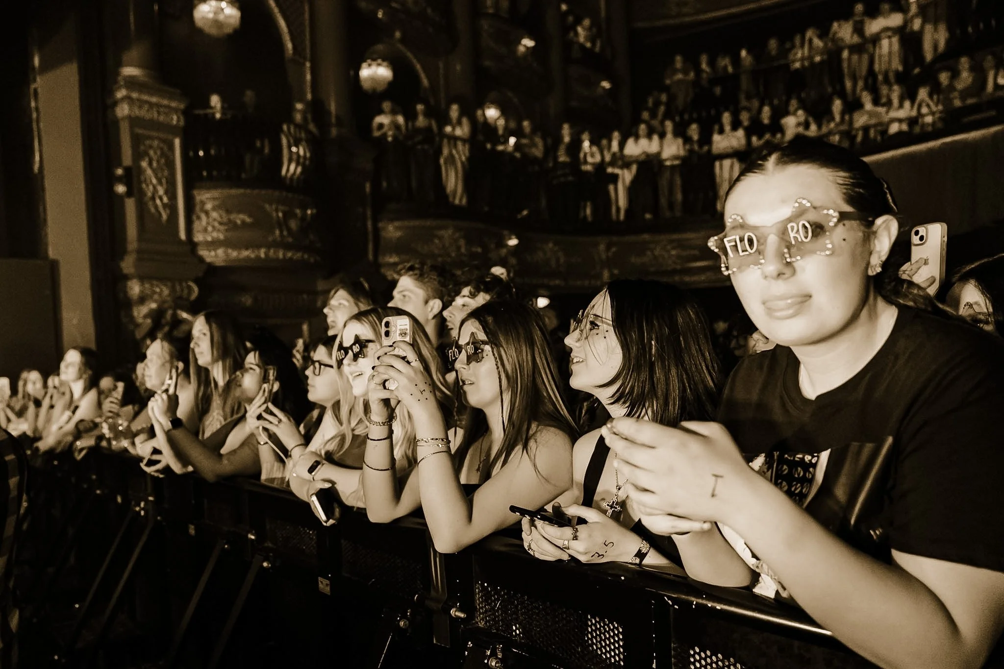 A crowd of people sitting in an indoor venue, many holding smartphones, with one woman wearing novelty glasses that spell "FLO RO" and others with face paint, watching an event.