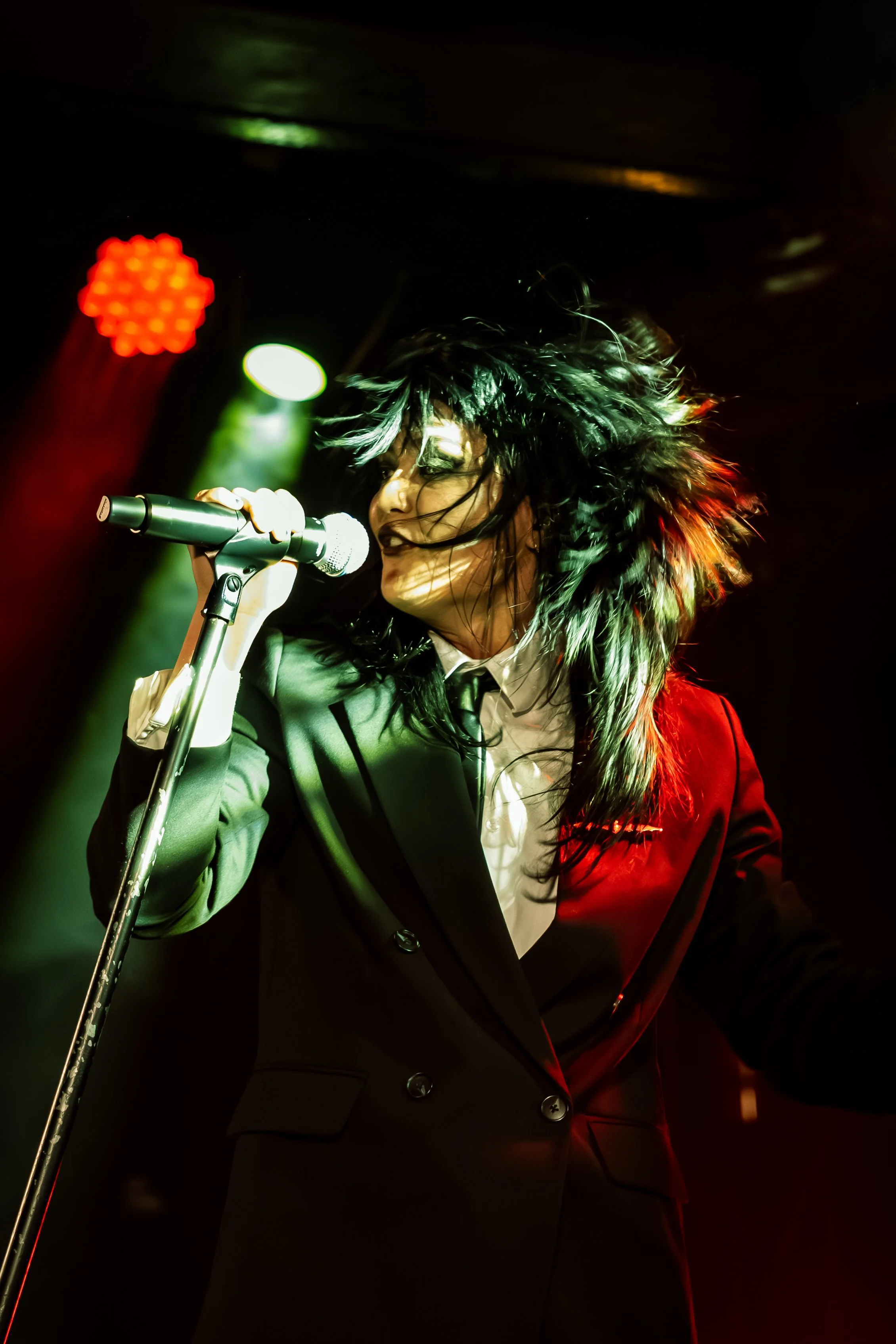 A person performing on stage, wearing a black suit with a white shirt, holding a microphone, with vibrant stage lighting and tousled black hair.
