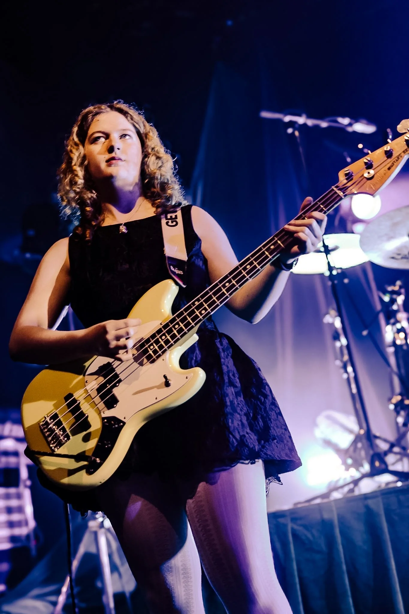 Young woman playing a yellow electric bass guitar on stage under purple and blue lights.