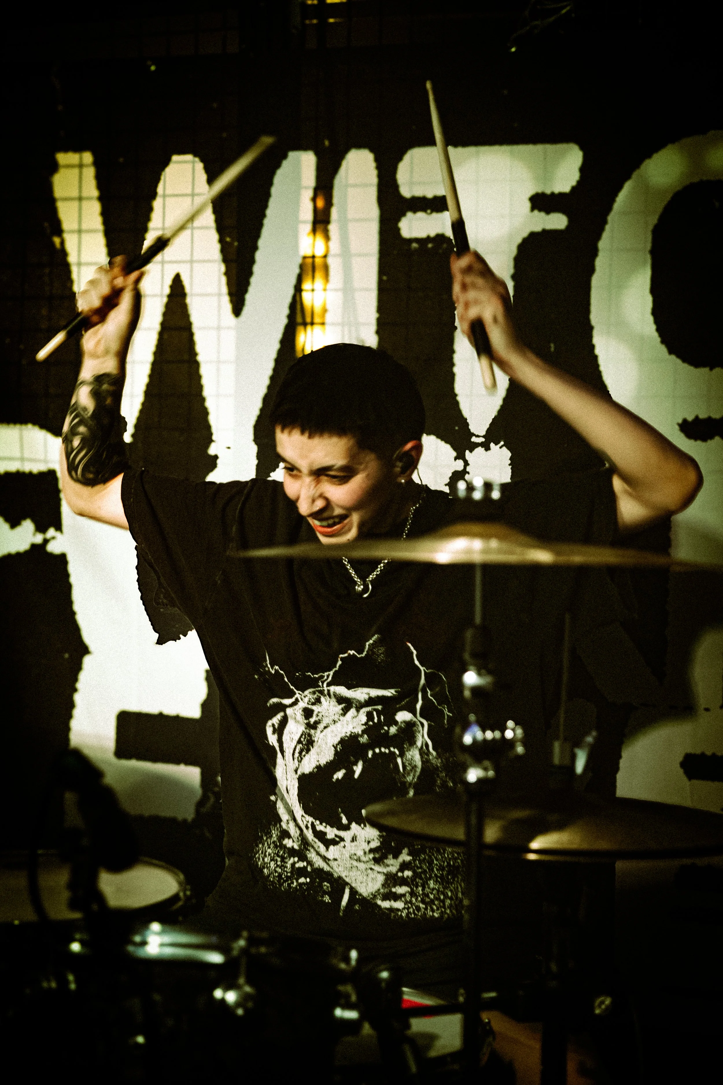 A person with short black hair playing drums on stage, wearing a black T-shirt with a large skull graphic. They are enthusiastically raising drumsticks, with a dark background featuring a large illuminated display.