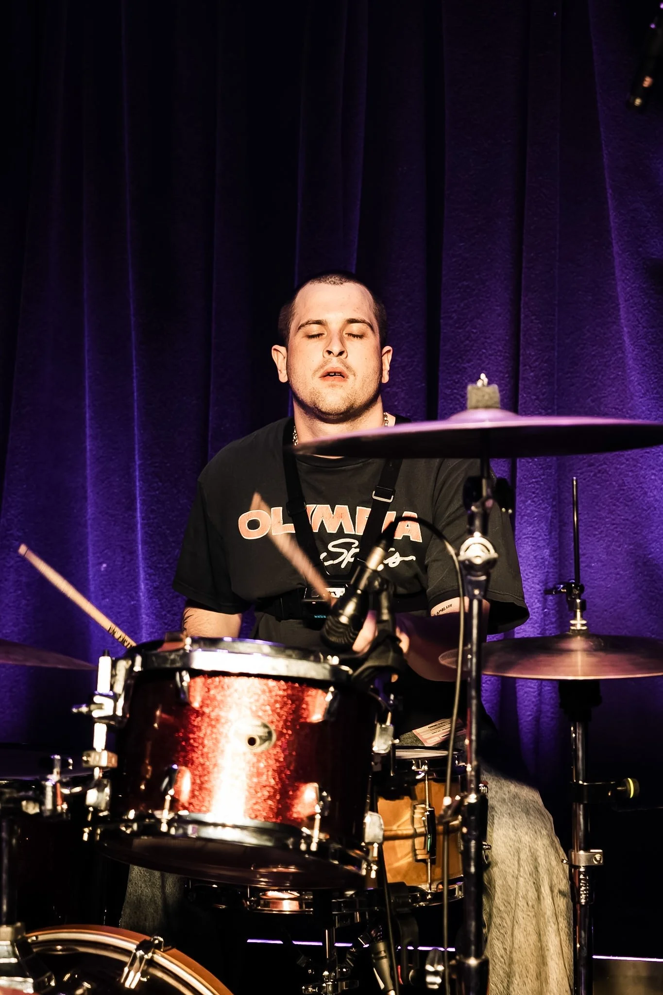 A man with short dark hair and a black t-shirt playing a drum set on stage with dark purple curtains in the background.