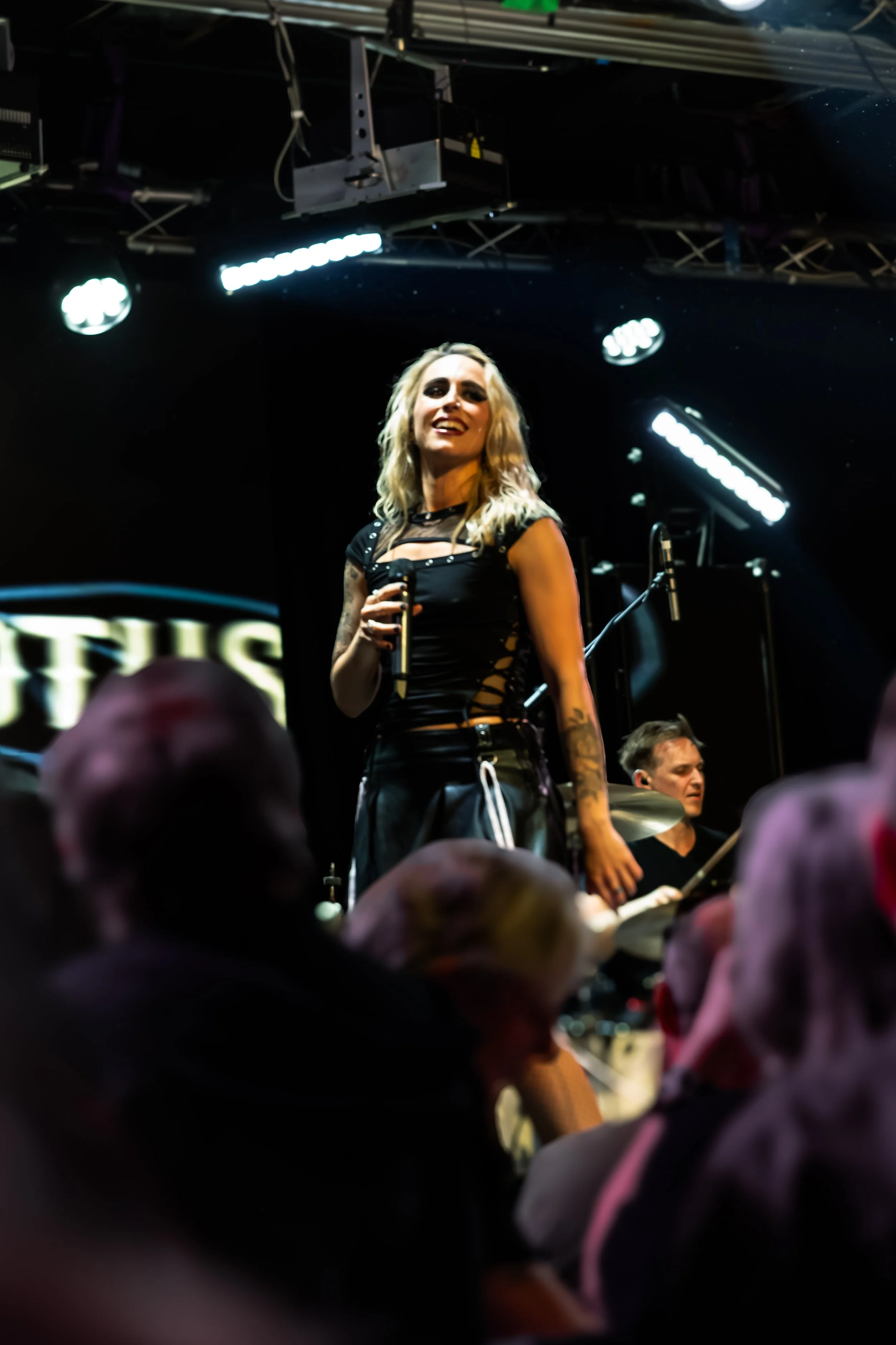 A woman with blonde hair and tattoos on her left arm, wearing a black outfit, stands on stage holding a microphone, with an orchestra and stage lighting in the background. The audience is visible in the foreground.