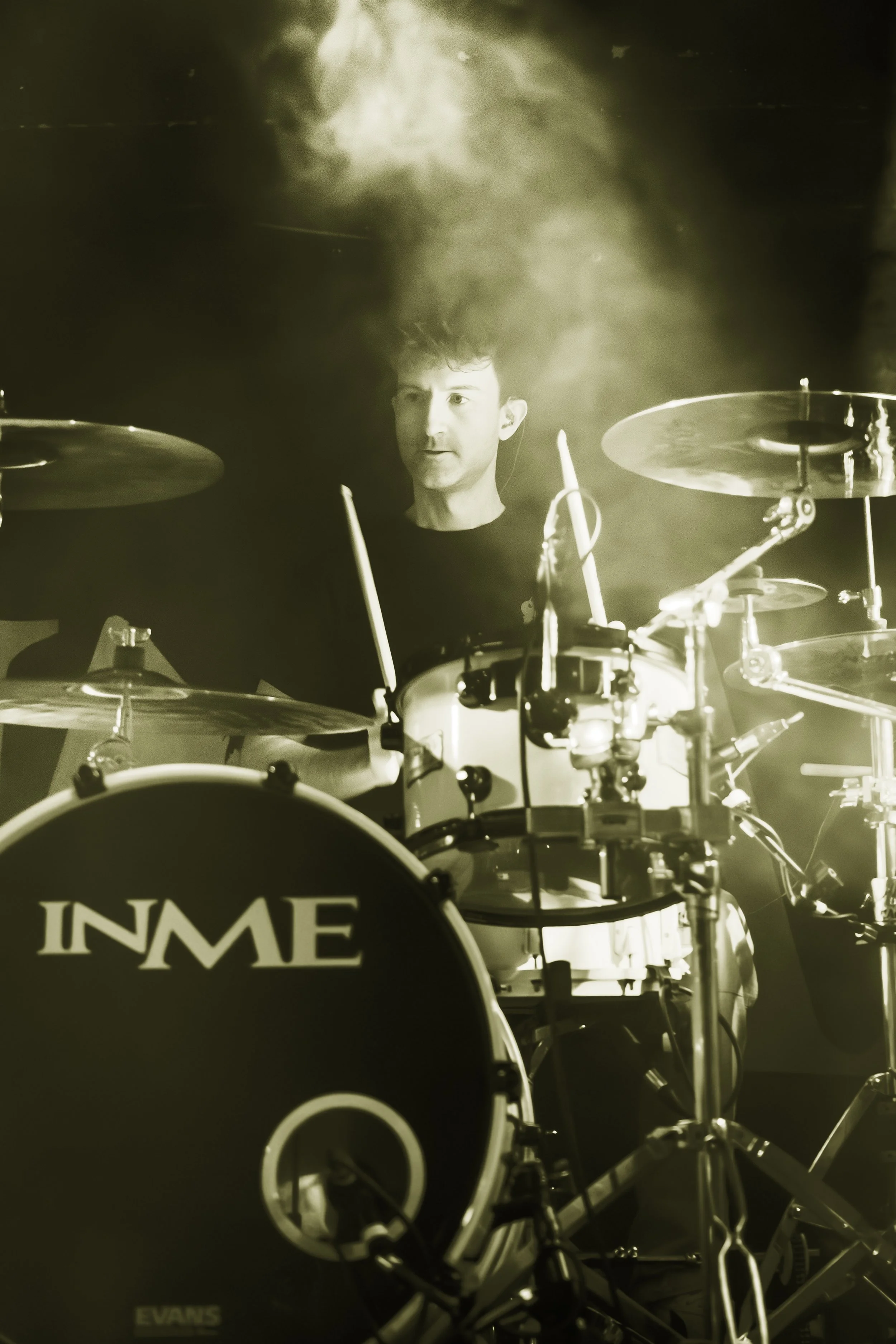 A black and white photo of a man playing the drums on stage, with focused expression, surrounded by drum equipment and smoke or fog.