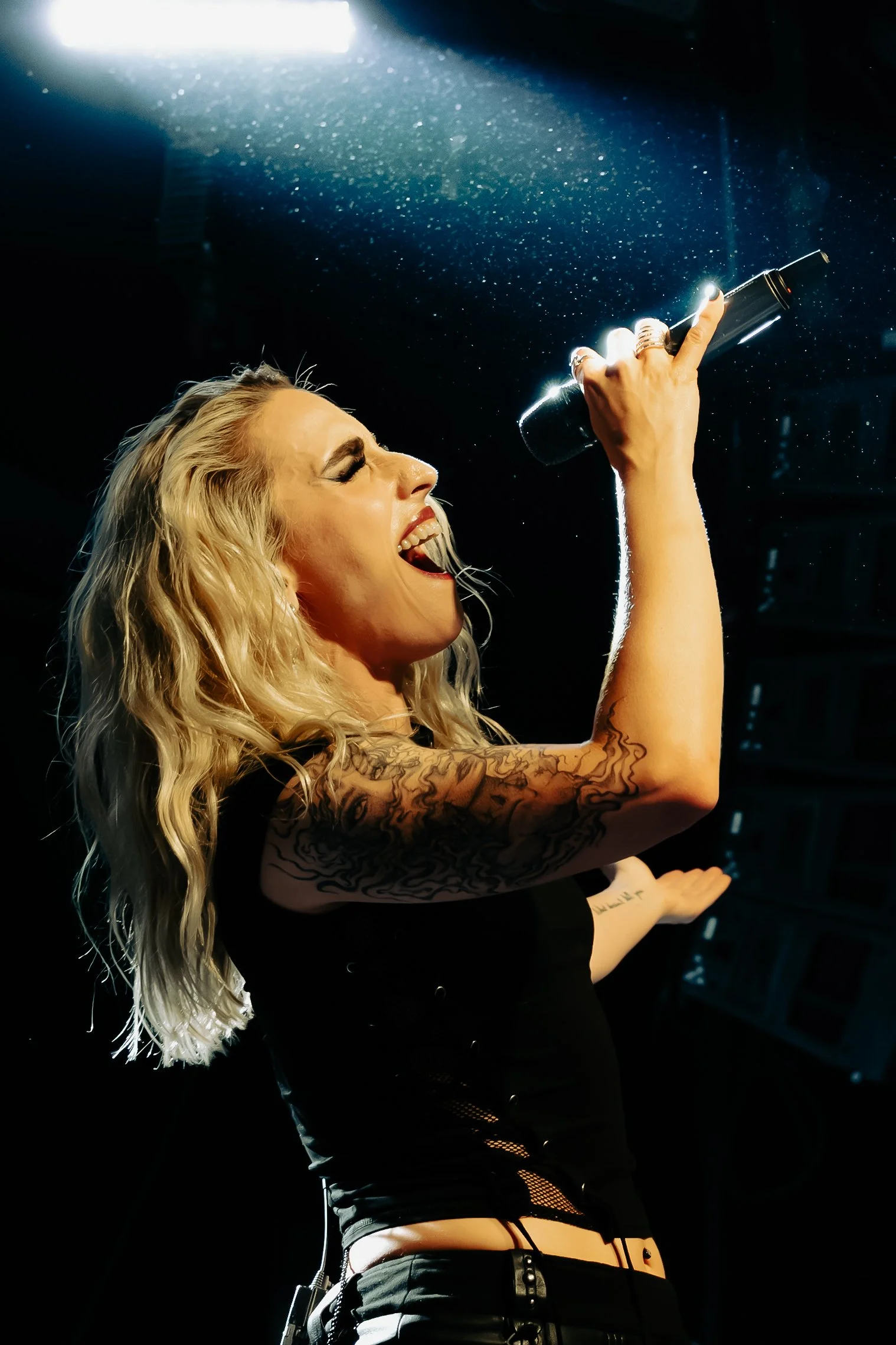 A female singer with blonde curly hair passionately singing into a microphone on a dimly lit stage, illuminated by a bright spotlight from above. She has tattoos on her arm and is wearing a black outfit with cutouts.