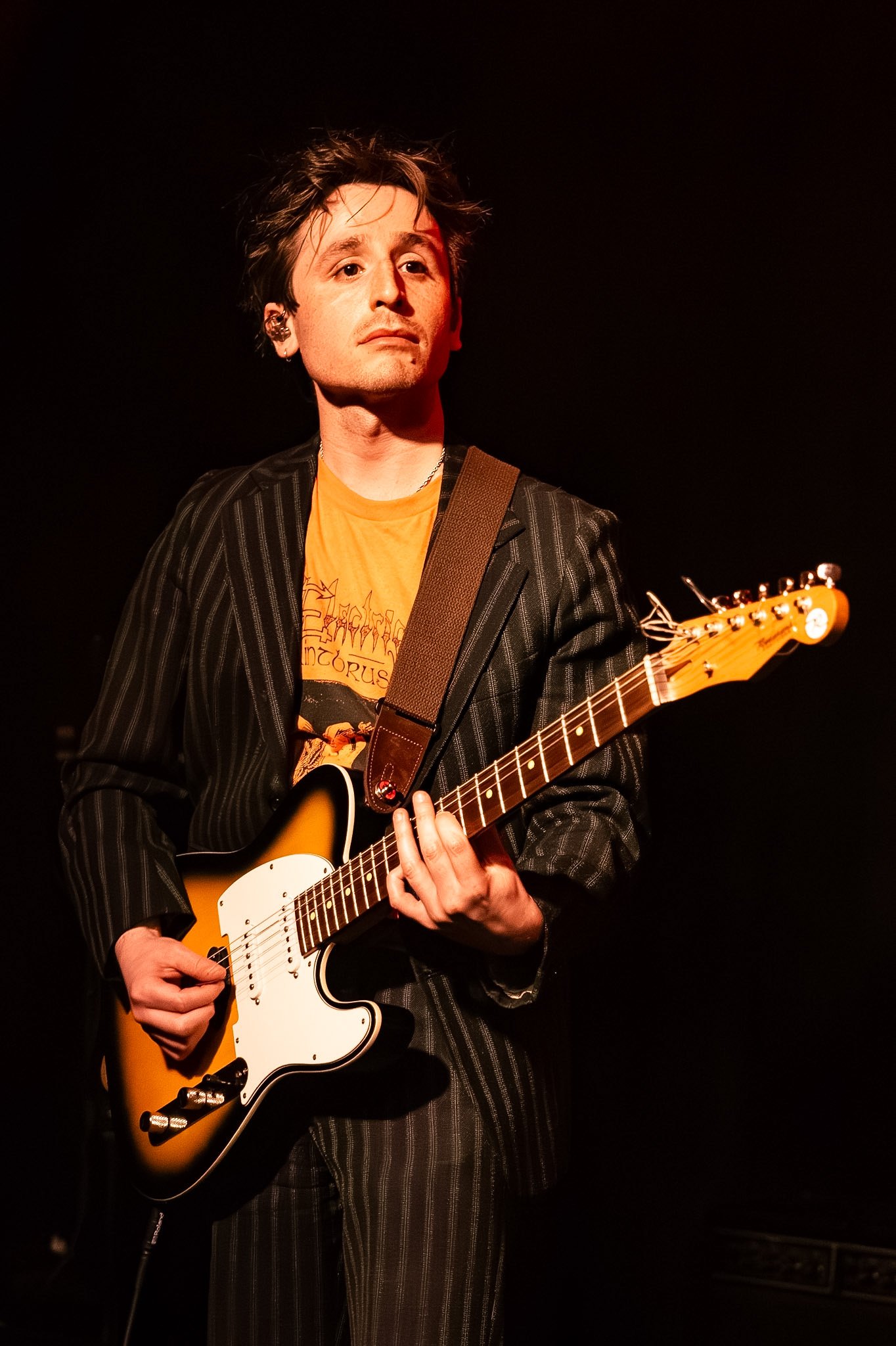 A male musician playing an electric guitar on stage, wearing a black pinstripe blazer and a yellow graphic T-shirt, with short, messy hair, under dark stage lighting.