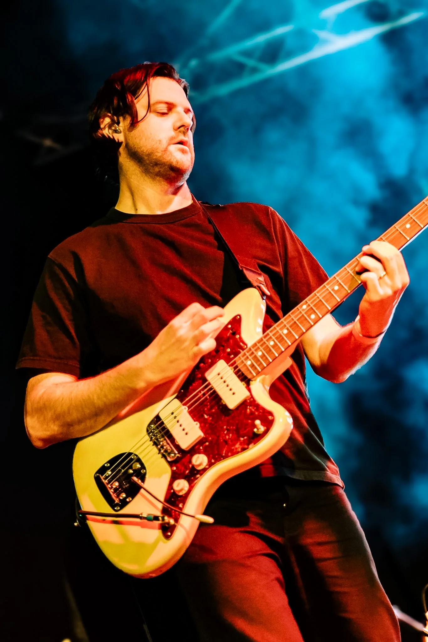A male musician with shoulder-length dark hair playing a cream-colored electric guitar on stage, illuminated by colored stage lights.