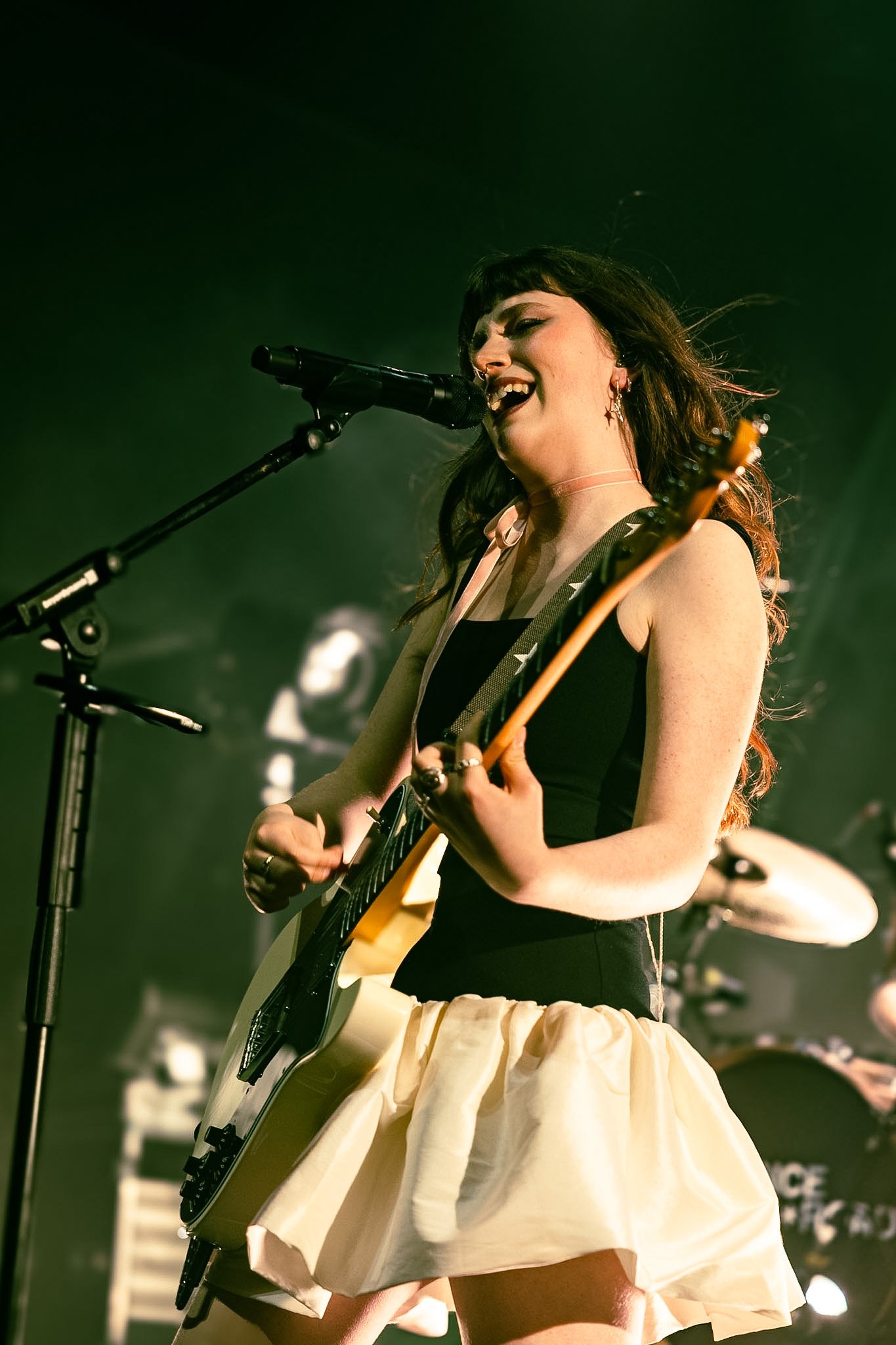 A female musician performing with an electric guitar and singing into a microphone during a concert, with a backup drummer in the background.