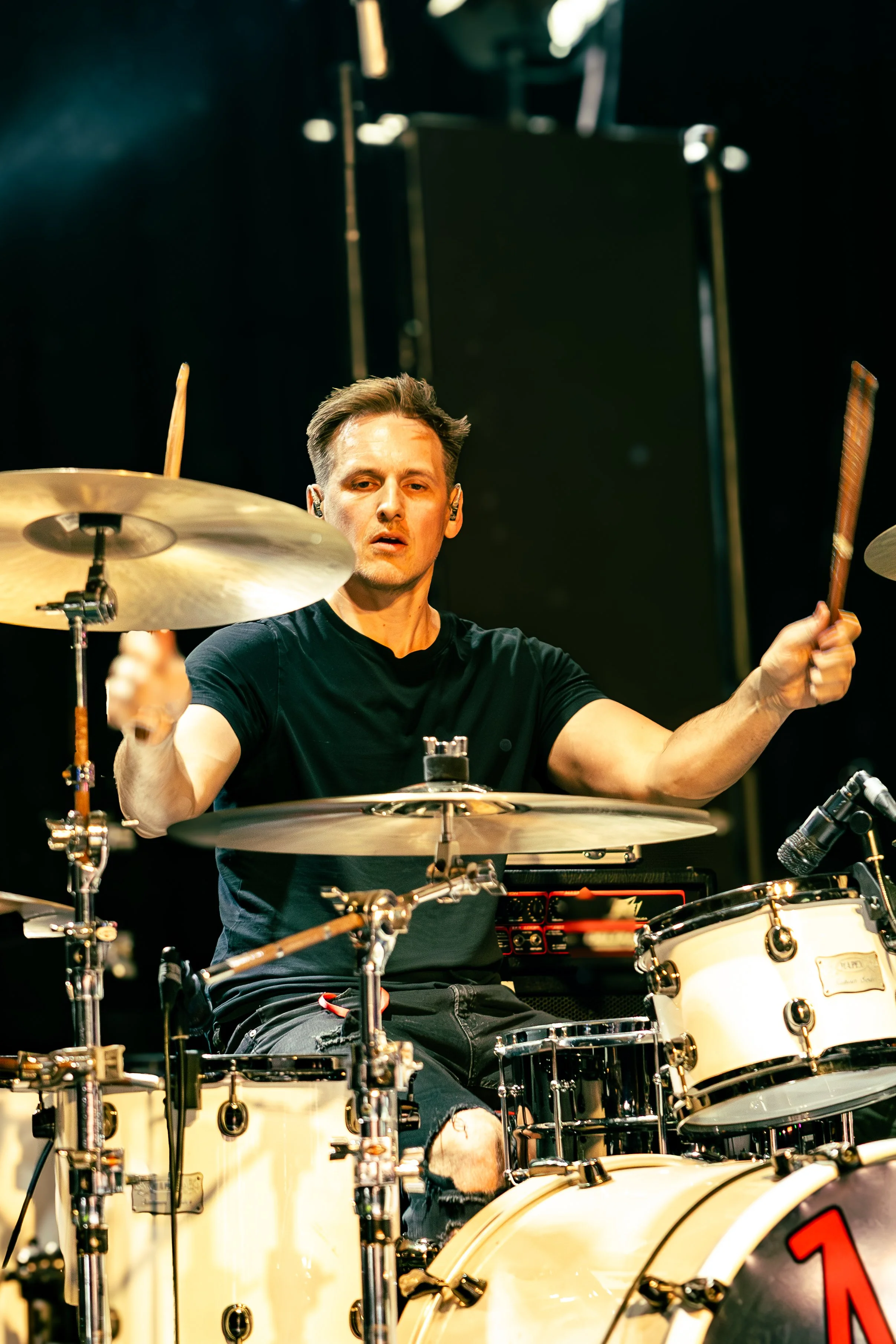 A man playing drums on stage with a focused expression. He is wearing a black T-shirt and sitting behind a white drum set. The background is dark, with stage lights highlighting him.