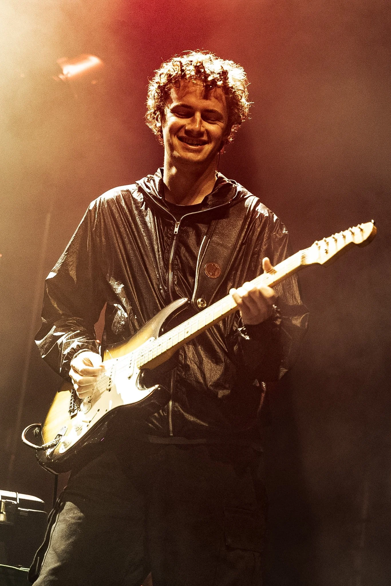 A man with curly hair smiling while playing an electric guitar on stage during a performance, wearing a shiny black jacket.