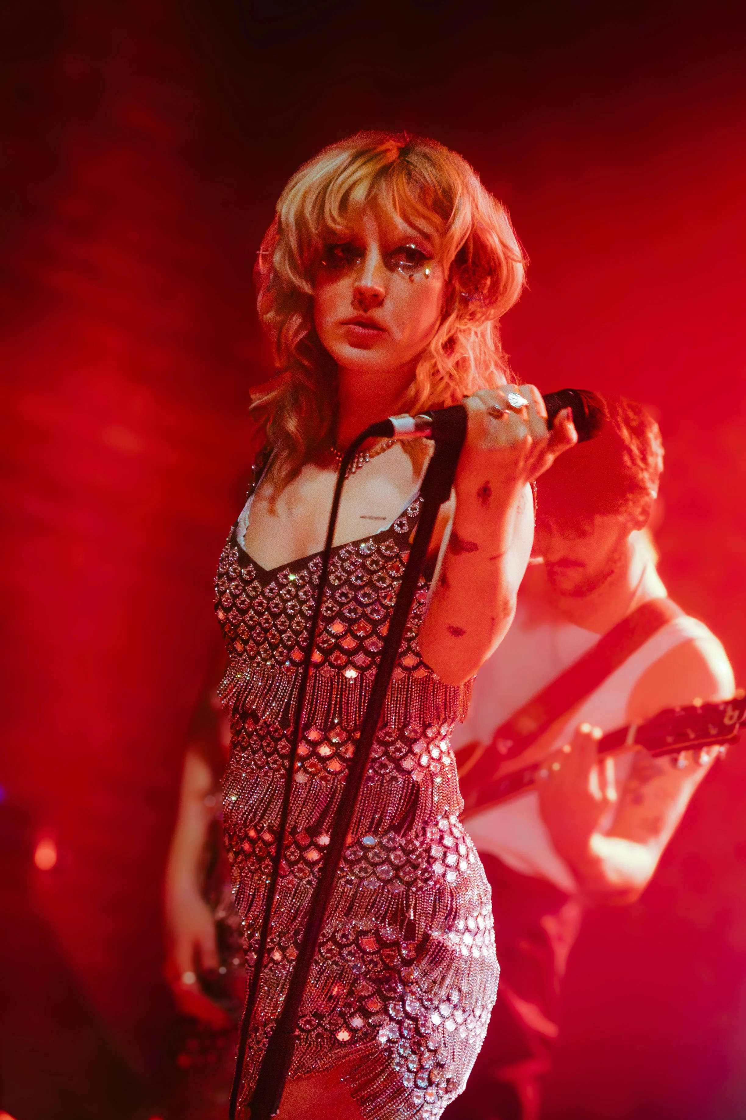 A woman with wavy blonde hair and sparkling eye makeup is holding a microphone. She is wearing a shimmering, form-fitting dress adorned with shiny embellishments, and the background is bathed in red lighting.