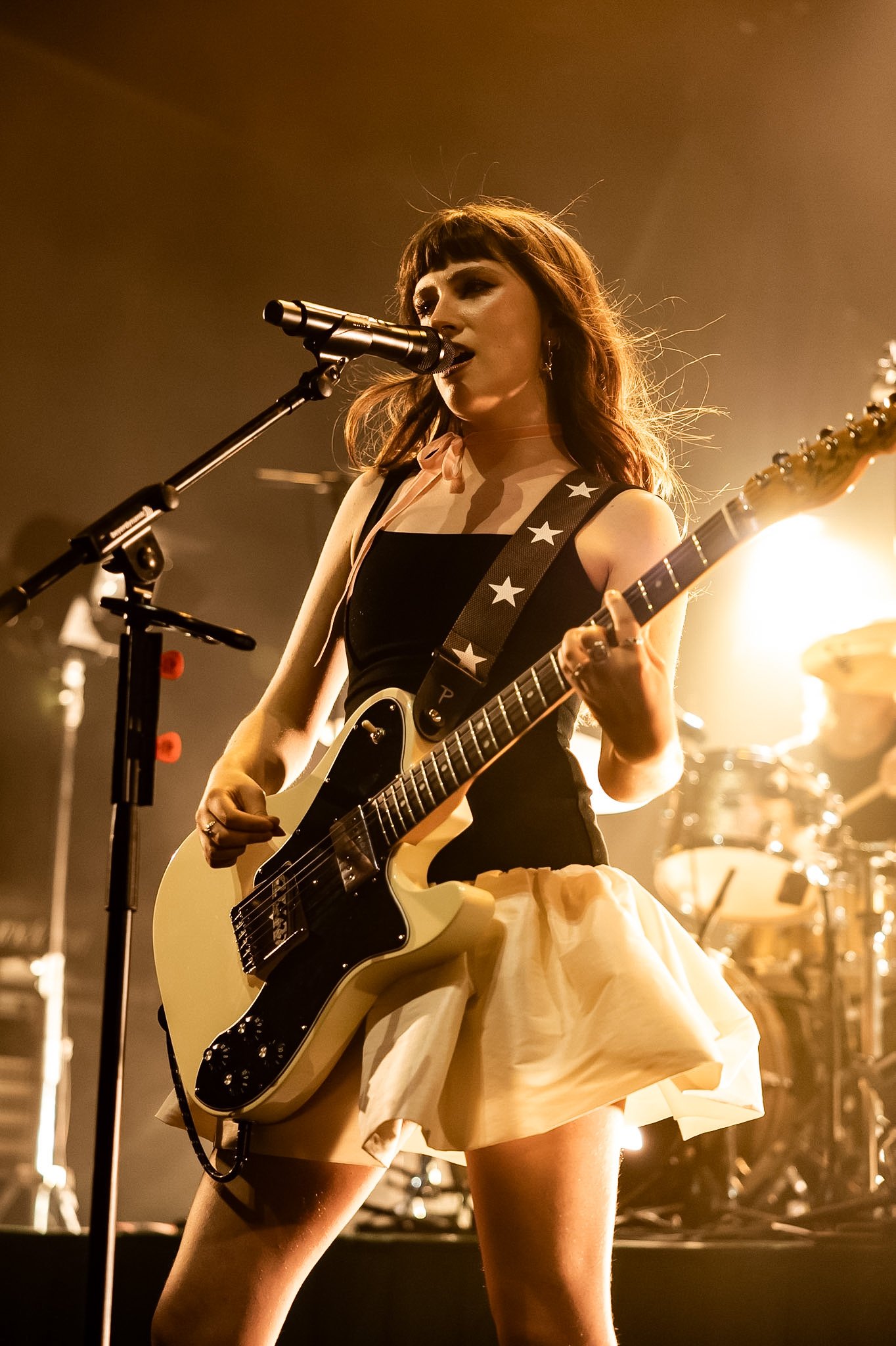 A female musician performing on stage with a yellow electric guitar, wearing a black top with star-patterned straps and a yellow skirt, under warm stage lighting.