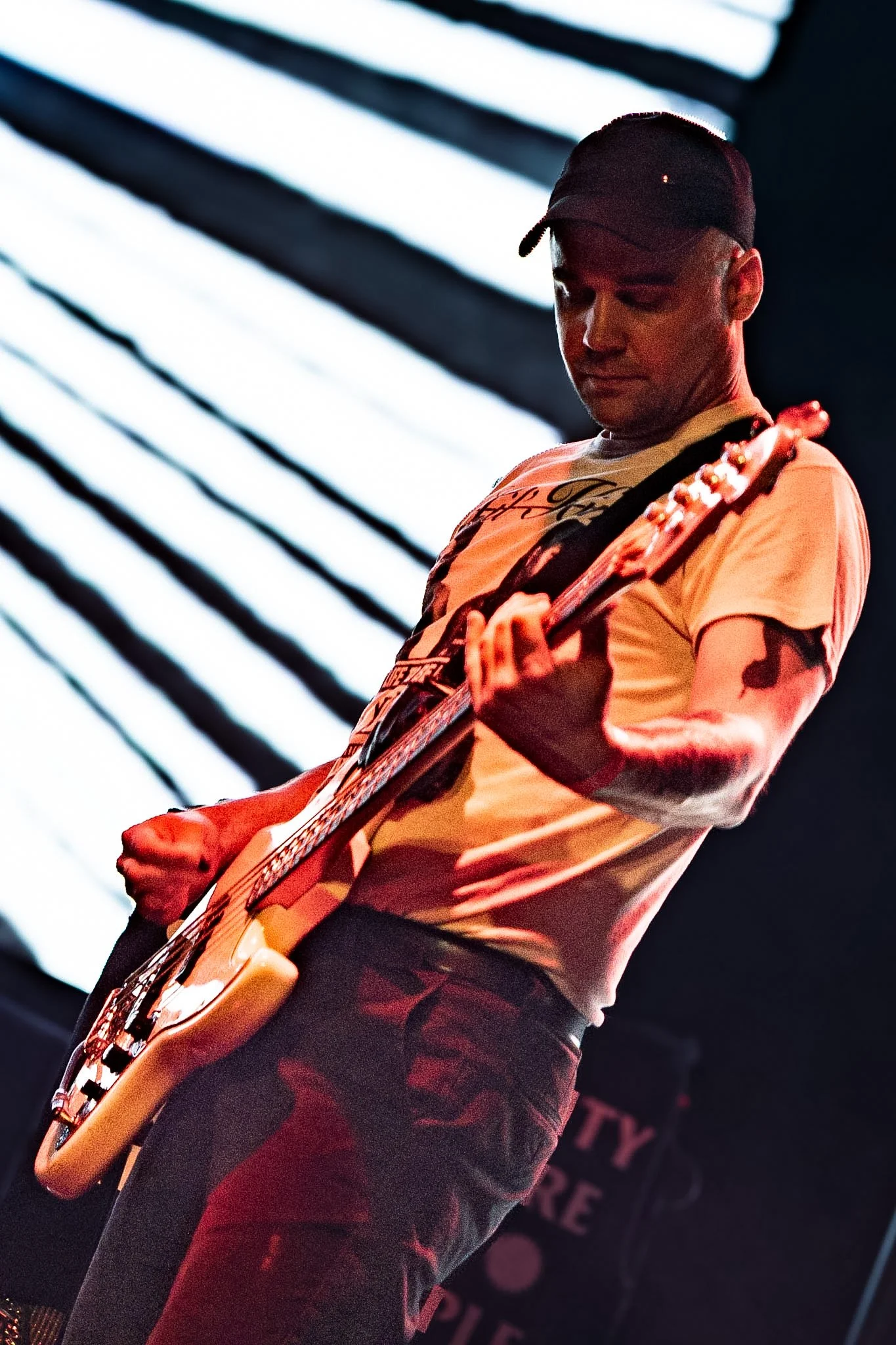 A person wearing a black cap and a light-colored t-shirt playing an electric guitar on stage, with a black background and bright stage lights.