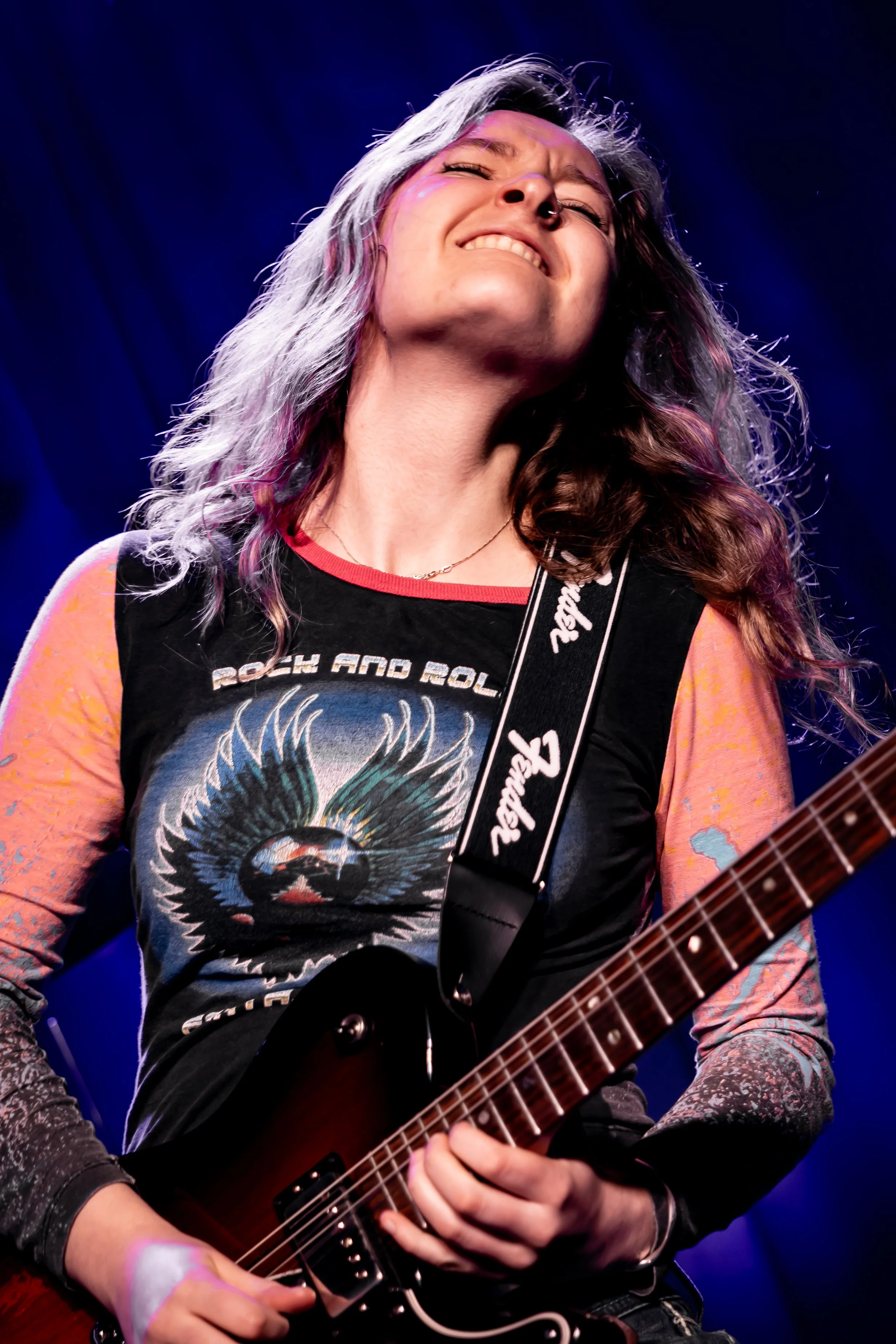 Person playing an electric guitar on stage, captured from the chest up, with a dark blue background. The person has long, wavy hair, and is wearing a graphic T-shirt and a necklace.