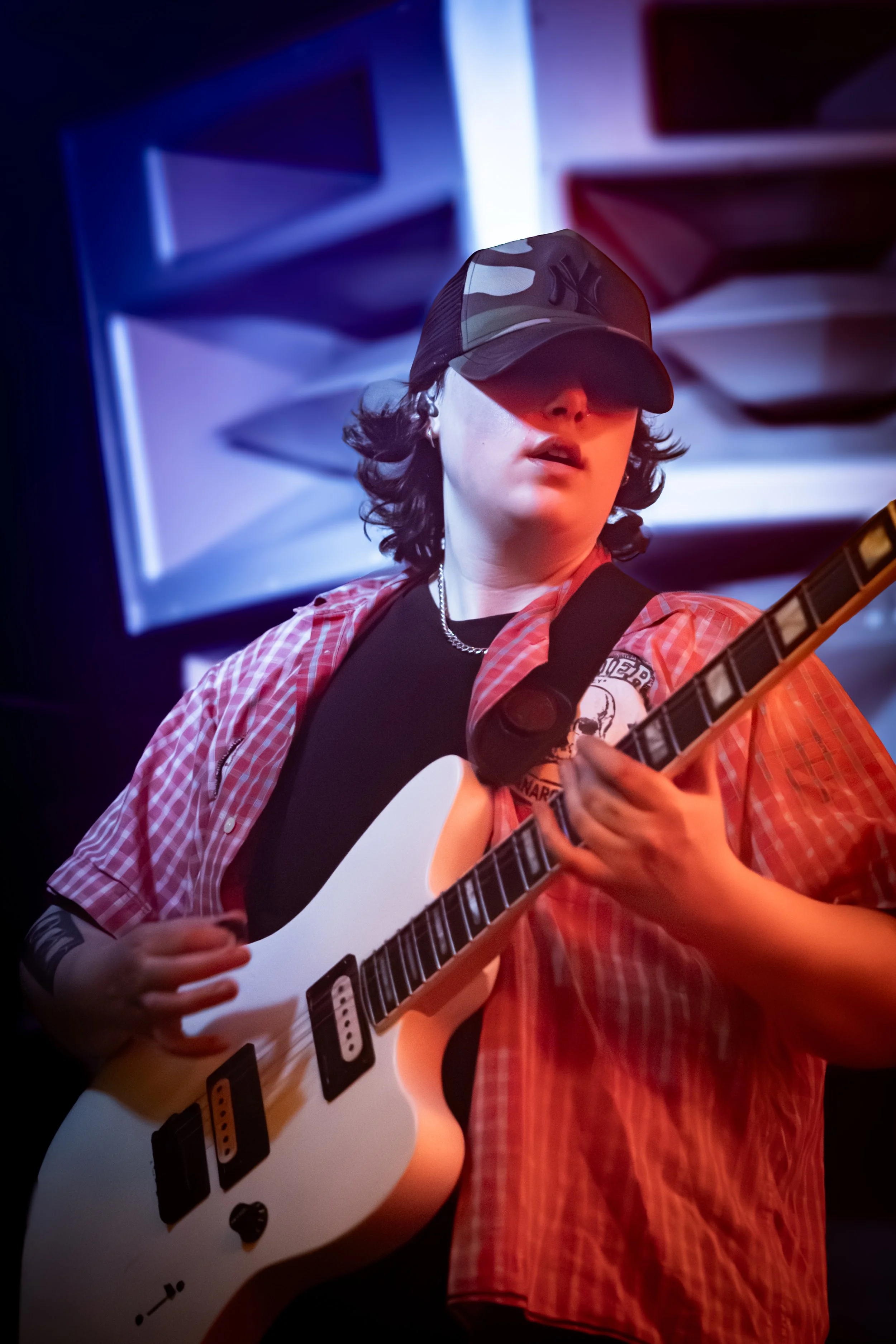 A young person with dark hair and a black cap wearing a red plaid shirt playing an electric guitar with a black strap, in a stage setting with colorful lighting.