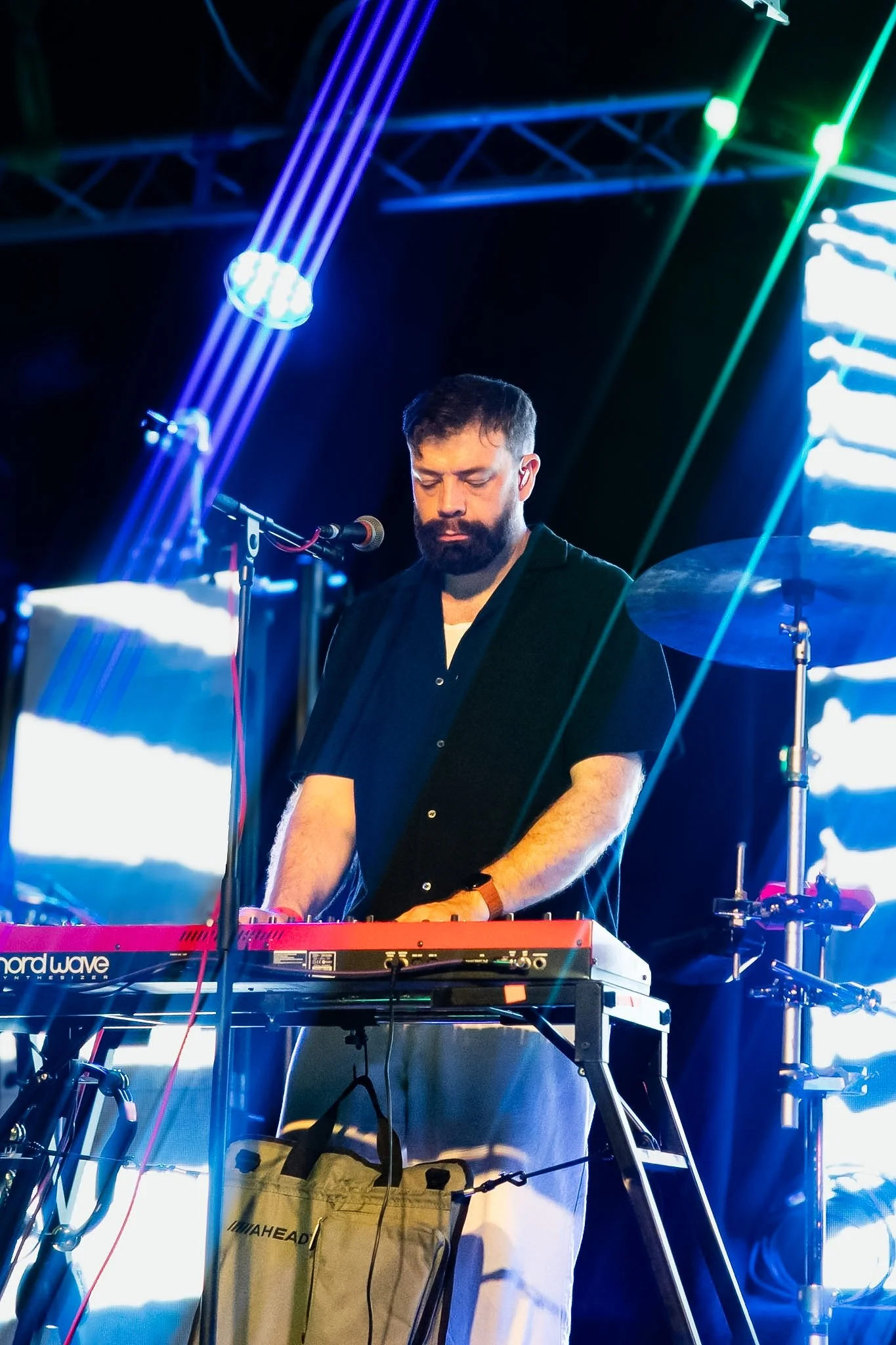A man with a beard and dark hair playing a red Nord Wave keyboard on stage with colorful laser lights and a drum set in the background.