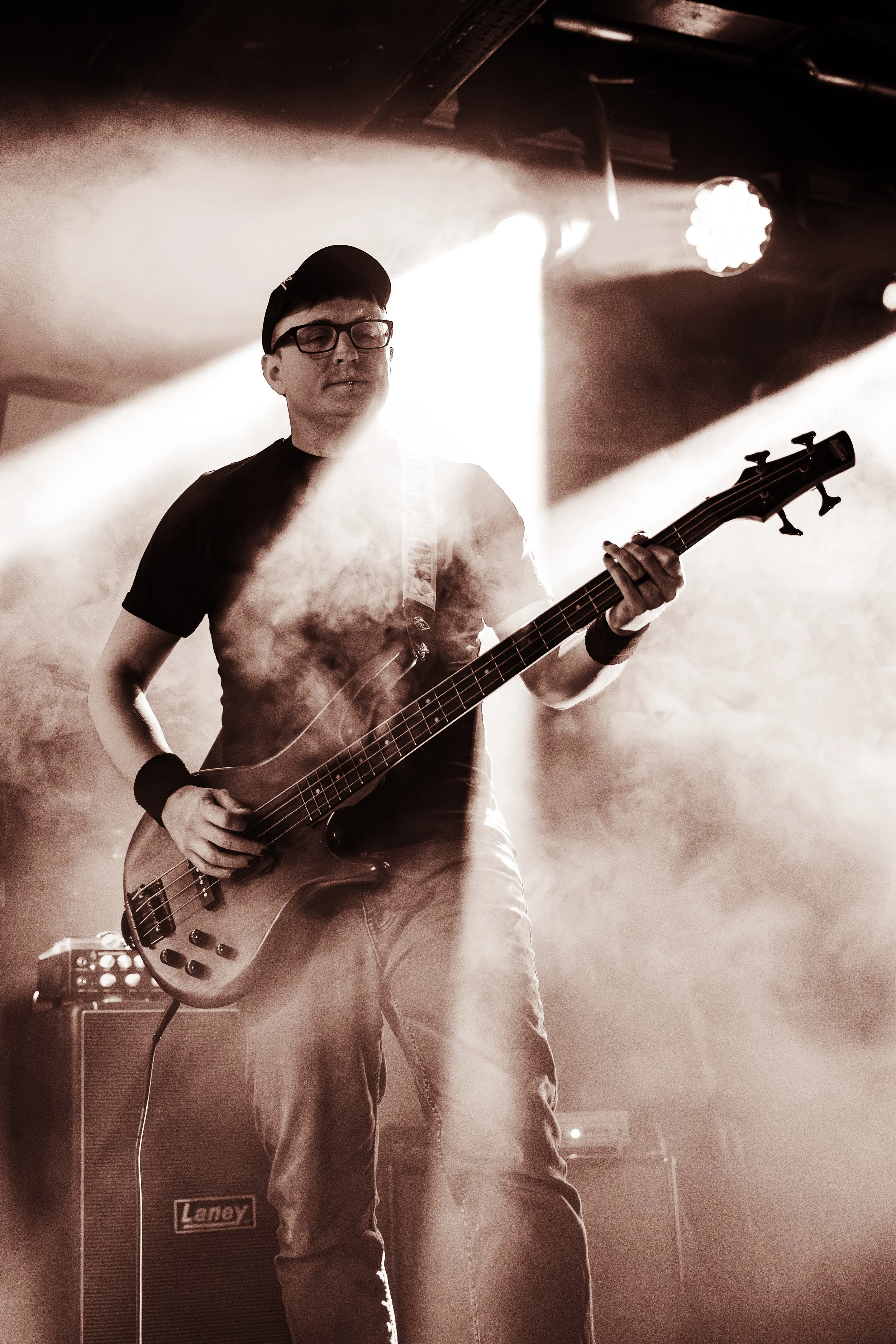 A musician playing an electric bass guitar on stage, illuminated by stage lights, with a Laney amplifier in the background and visible fog or smoke effects.