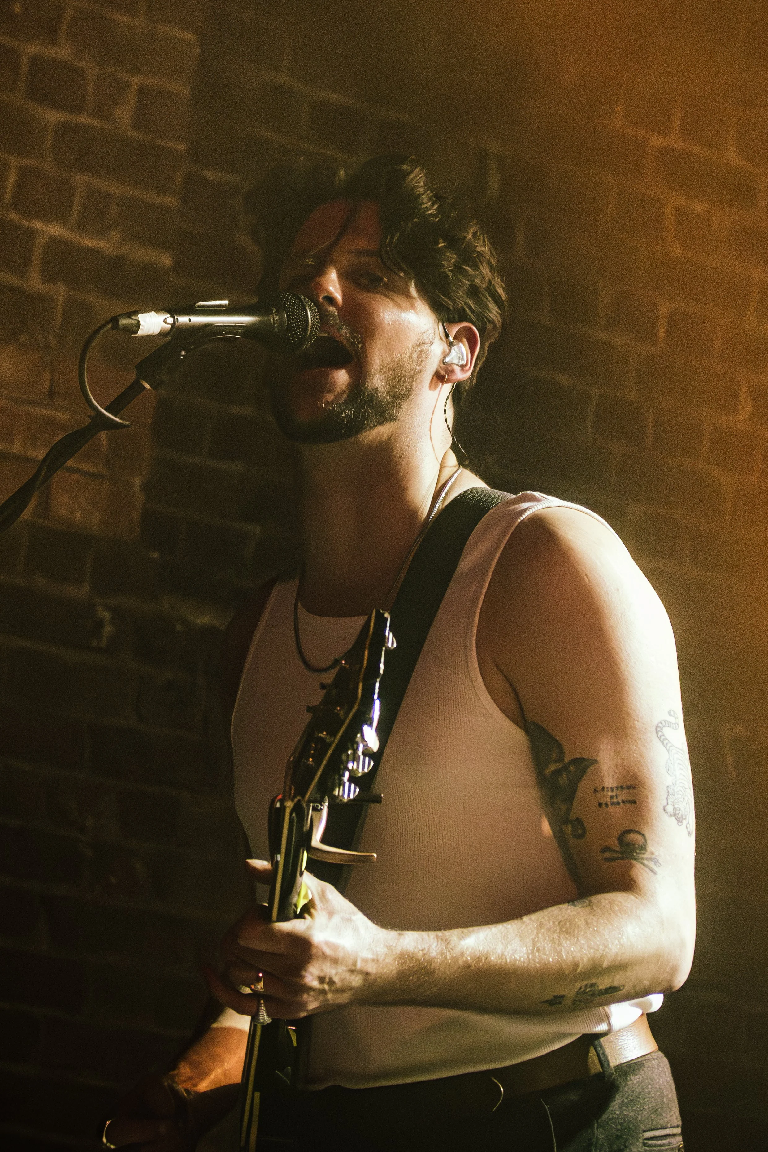 A male musician with dark hair, a beard, and tattoos, singing into a microphone while playing an electric guitar. He is wearing a white sleeveless shirt and has in-ear monitors. The background features a brick wall and warm lighting.