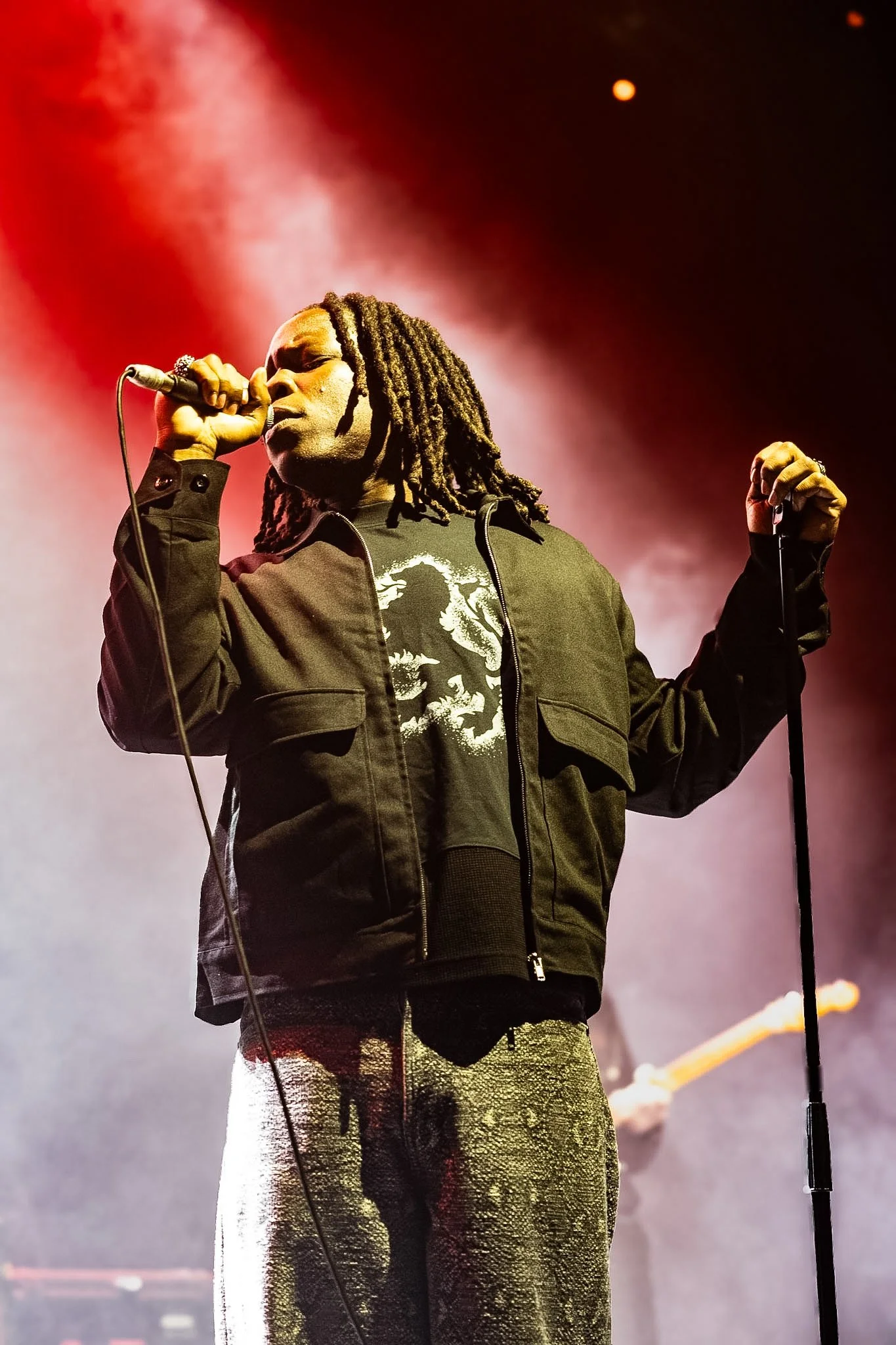 A person with dreadlocks singing into a microphone on stage with a dark background and orange lighting. Moio in concert at Koko Camden.
