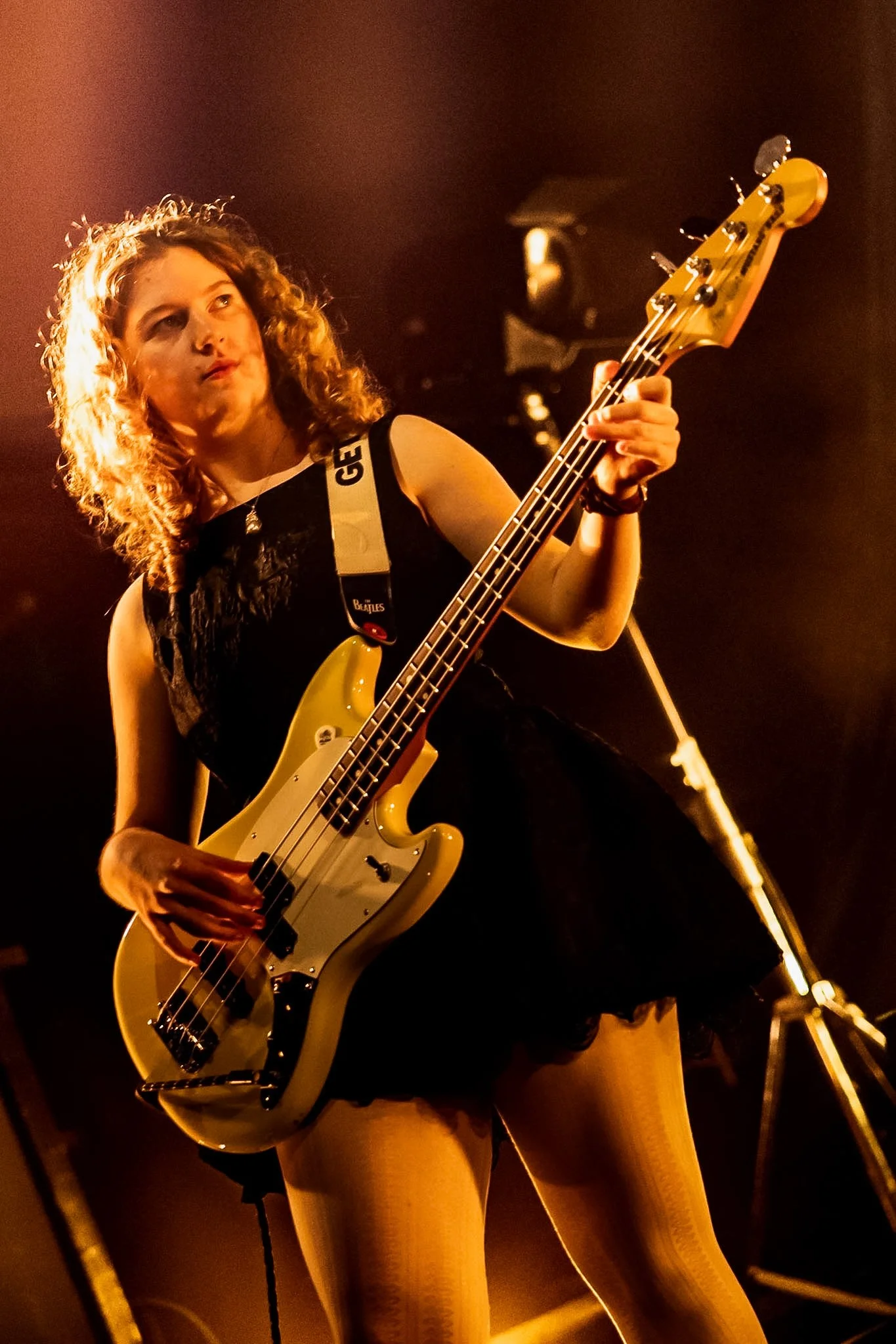 A woman with curly hair playing an electric bass guitar on stage, wearing a sleeveless top and bright yellow fishnet stockings, with stage lights illuminating her and a blurred microphone stand in the background.