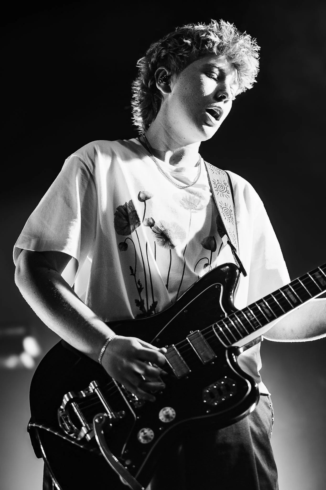 A woman with short, curly hair singing and playing an electric guitar. She is wearing a t-shirt with flower designs and a necklace. The photo is black and white.