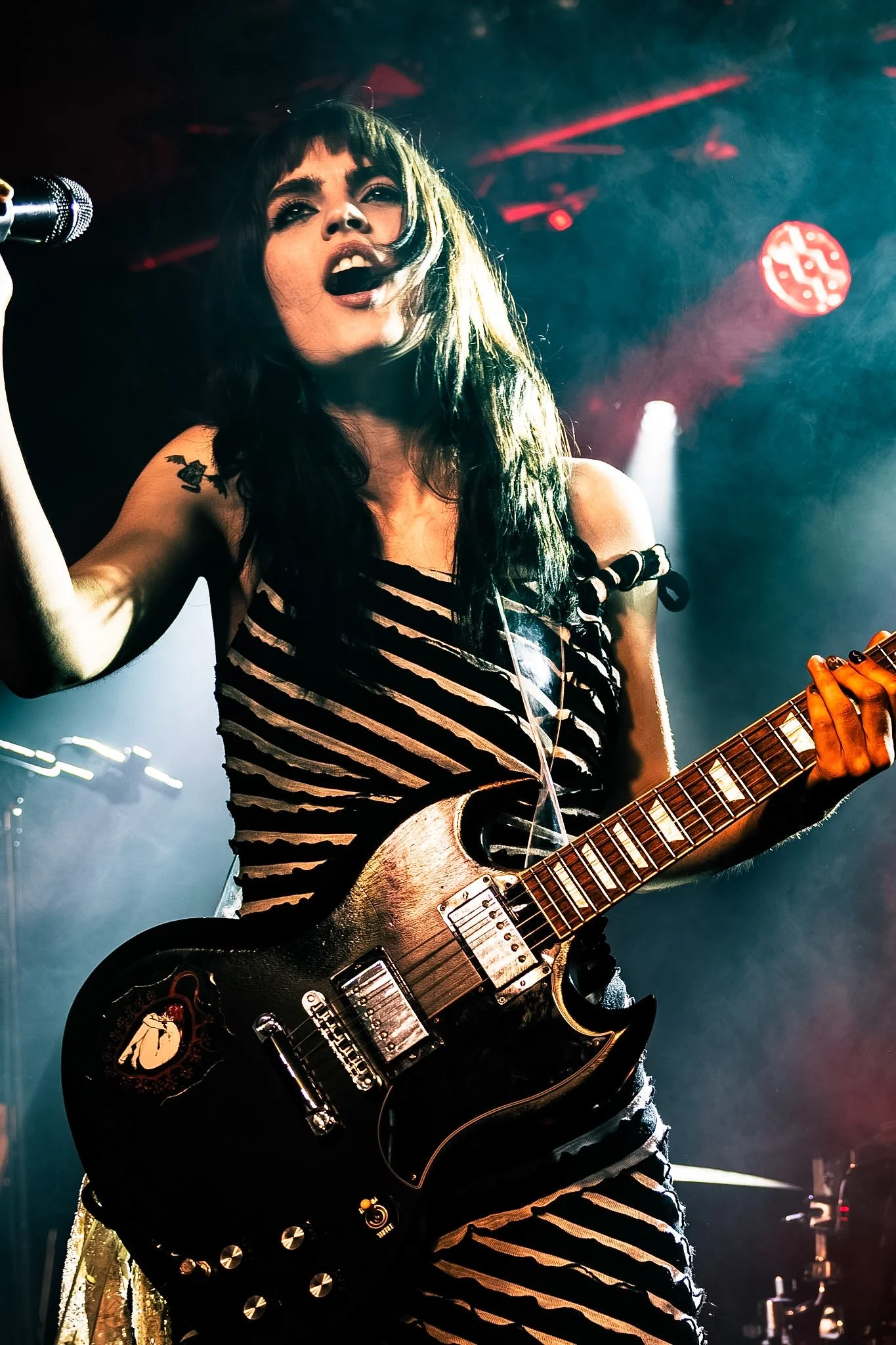 A female musician with dark hair and tattoos performs on stage, singing into a microphone while playing an electric guitar with stickers on it. The stage is dimly lit with colorful lights.