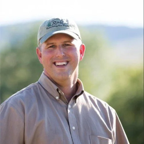 A man smiling outdoors wearing a beige button-up shirt and a baseball cap with a logo.